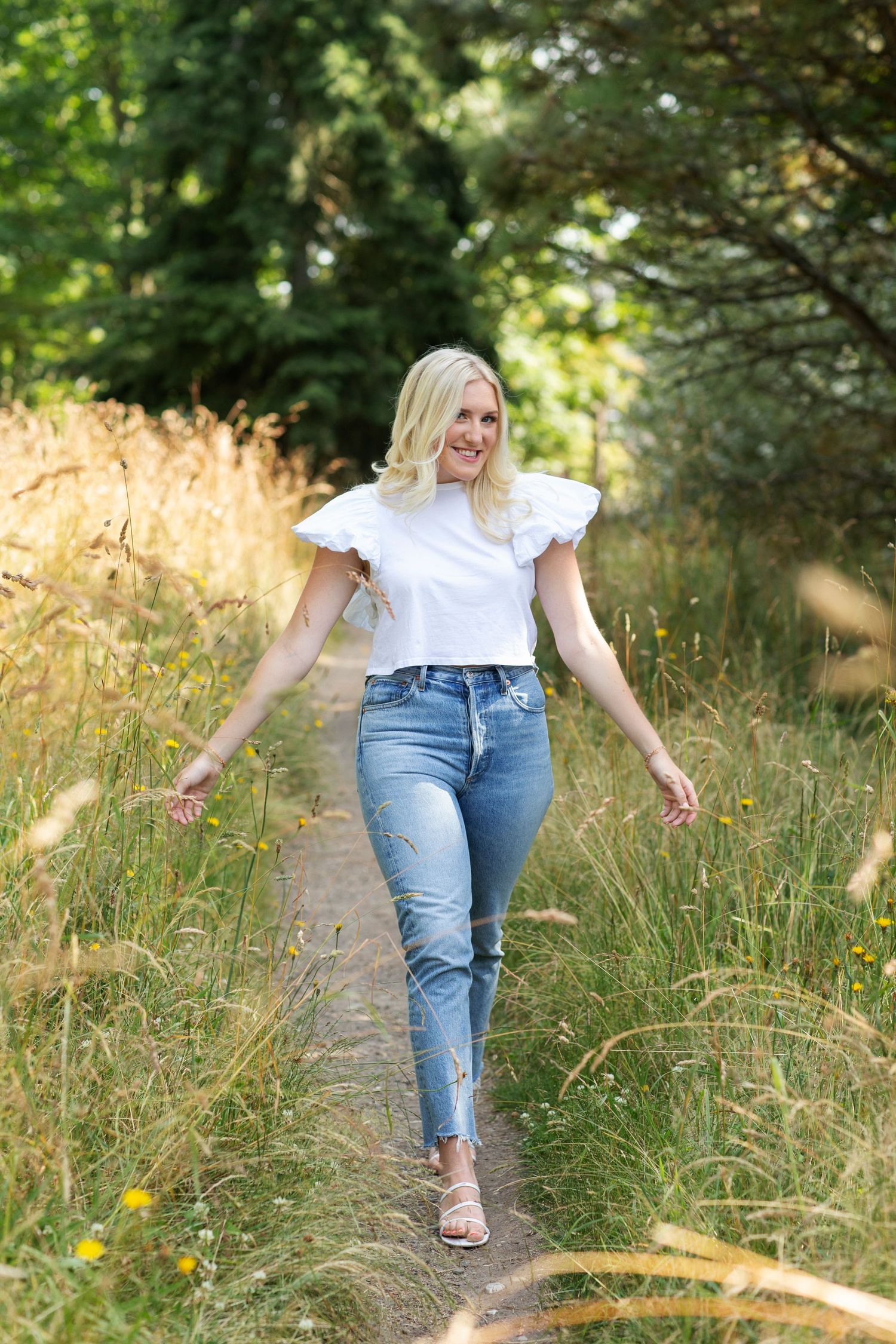 Blonde woman walking on a nature path wearing a white ruffle top and blue jeans surrounded by tall grass.