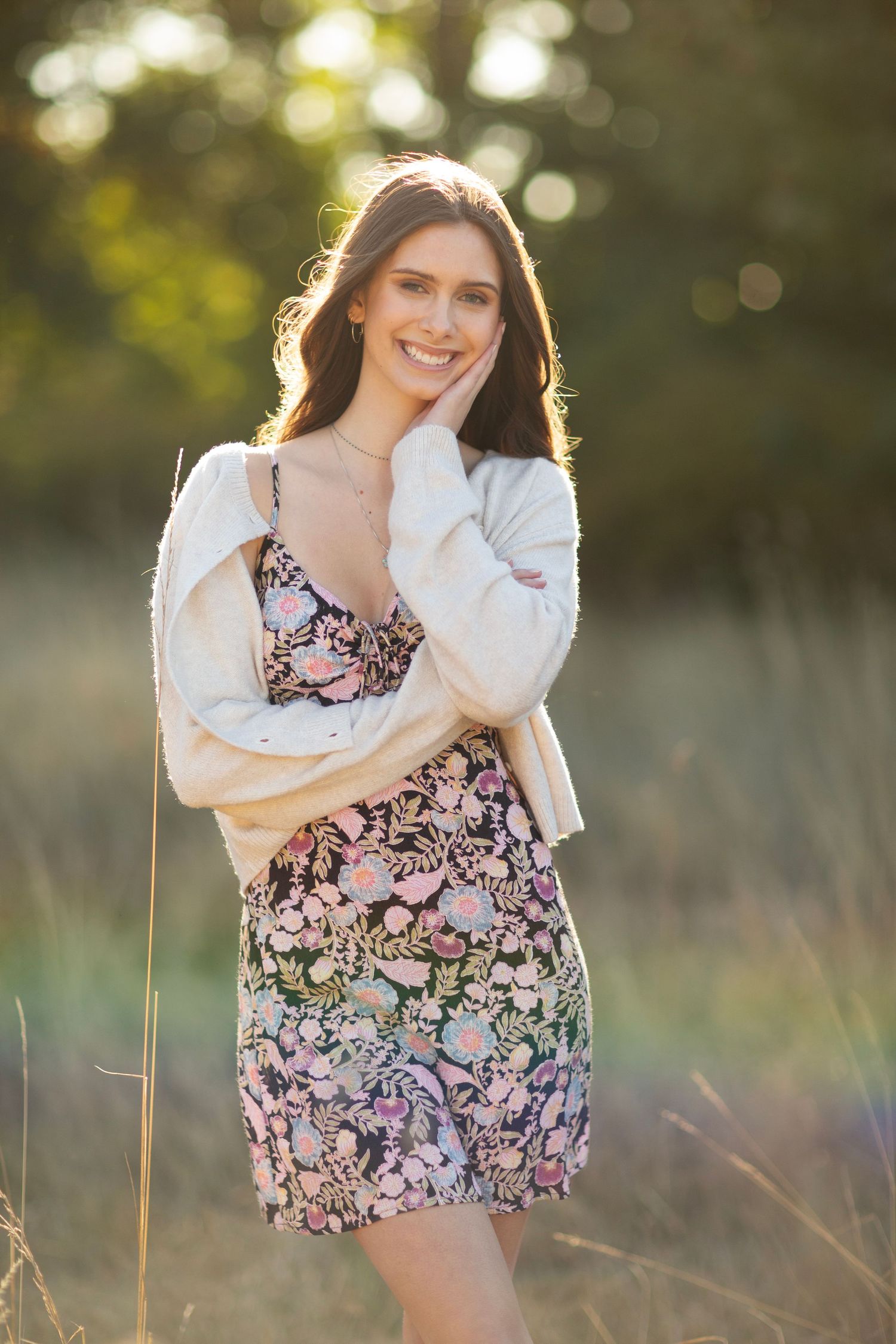 Smiling woman in floral dress and cream cardigan poses in warm sunlit outdoor grass field.