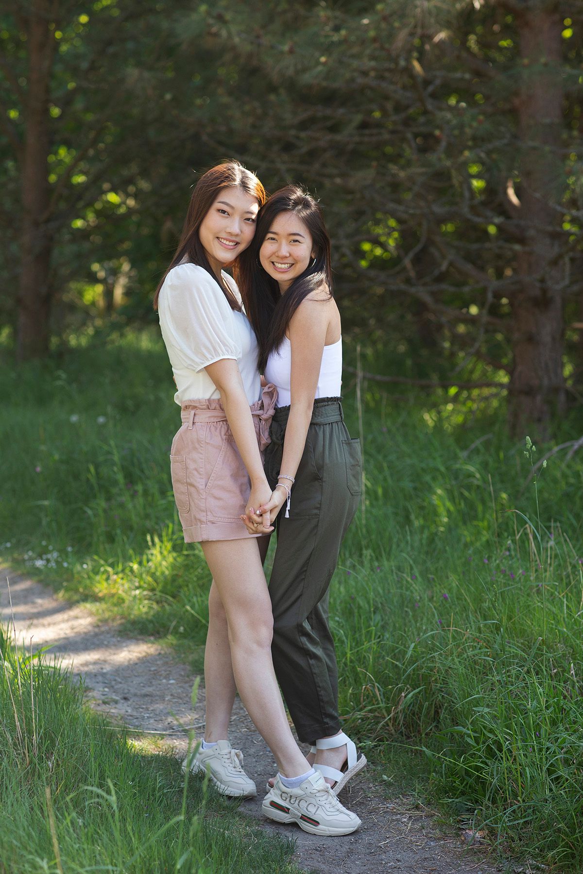 Two young Asian women holding hands and smiling on a forest path, wearing casual summer outfits.
