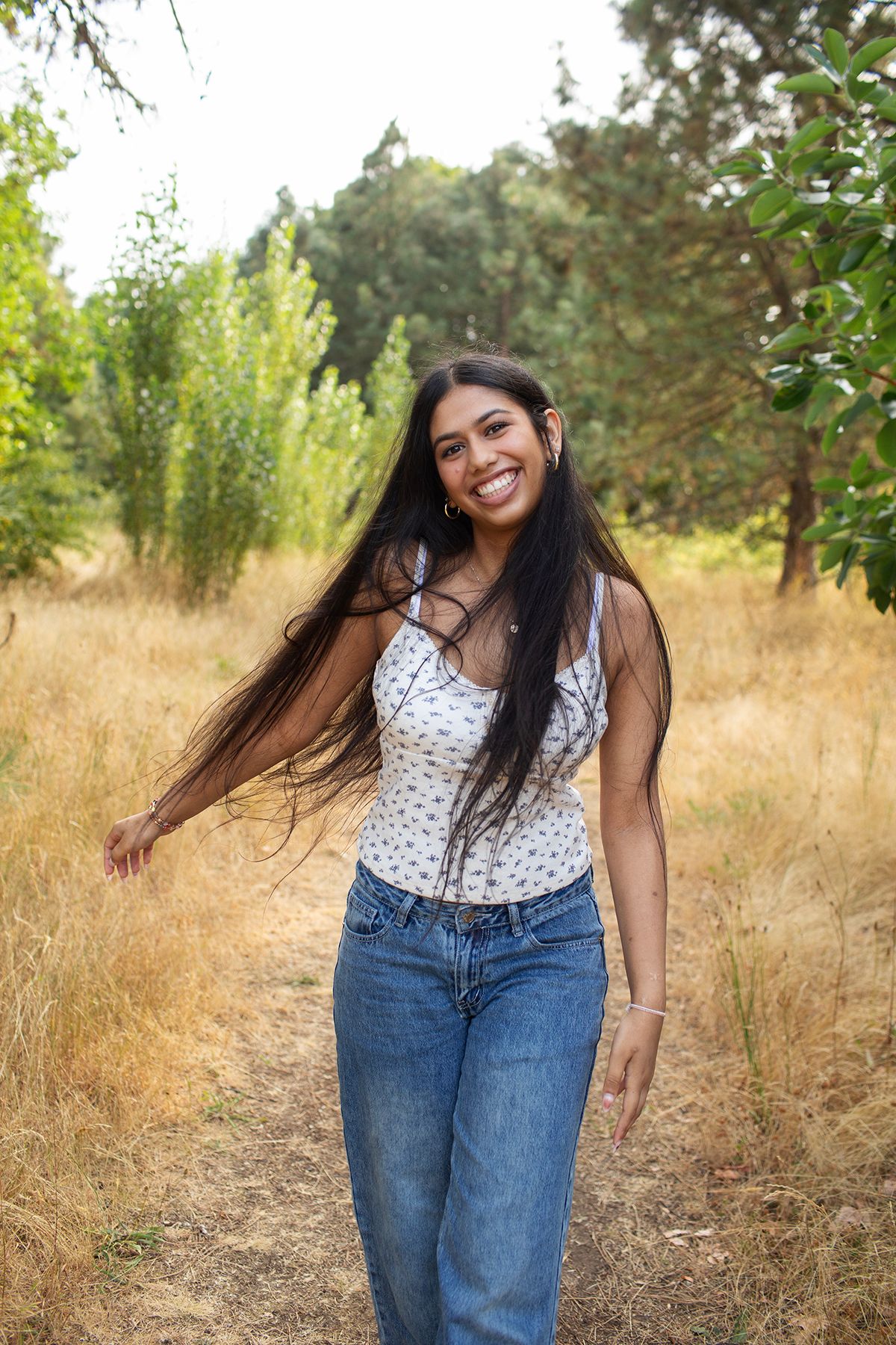 Smiling young woman with flowing dark hair walking along a golden grass path through green trees.