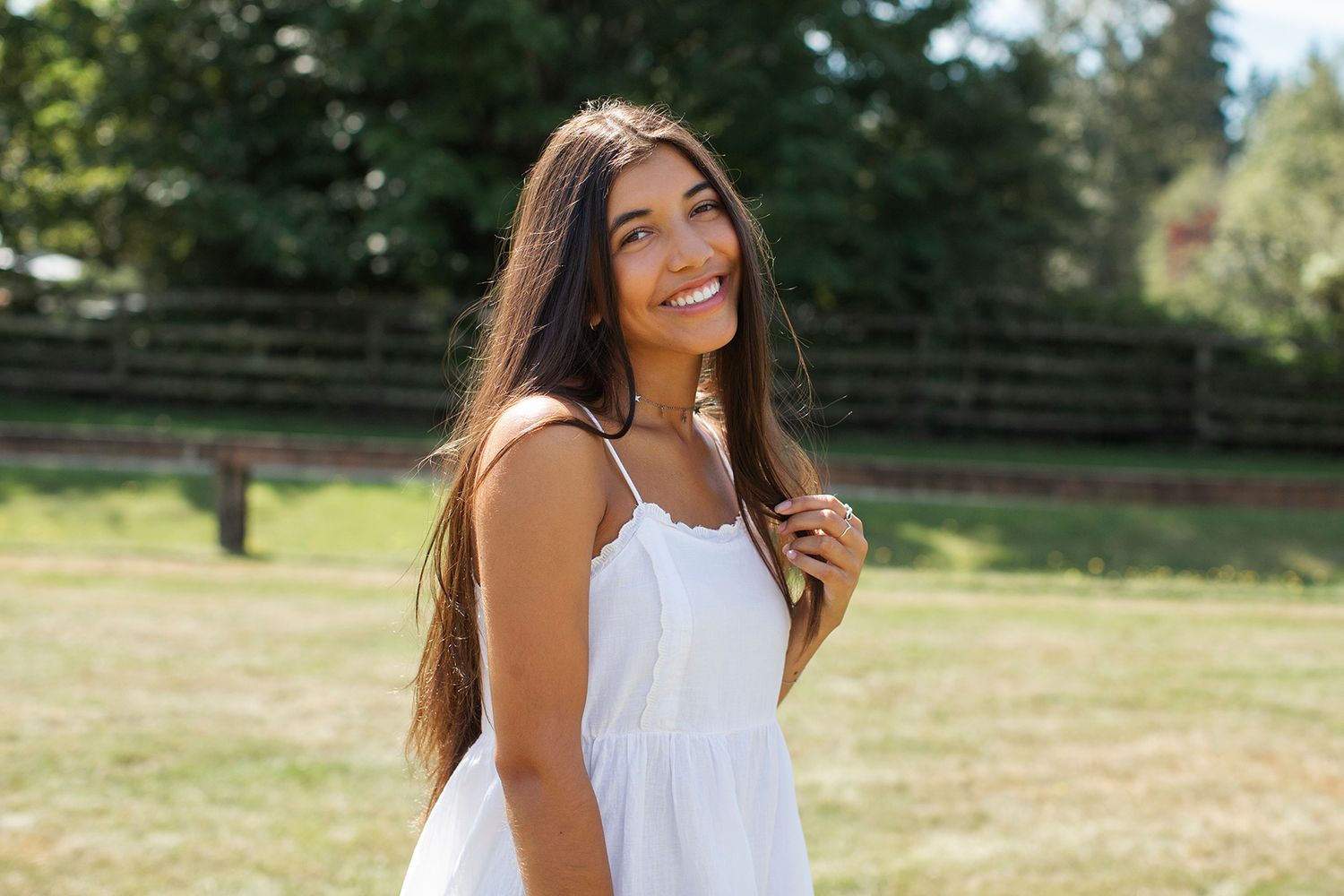 Smiling young woman with long dark hair wearing a white sundress standing outdoors in a sunny green field.
