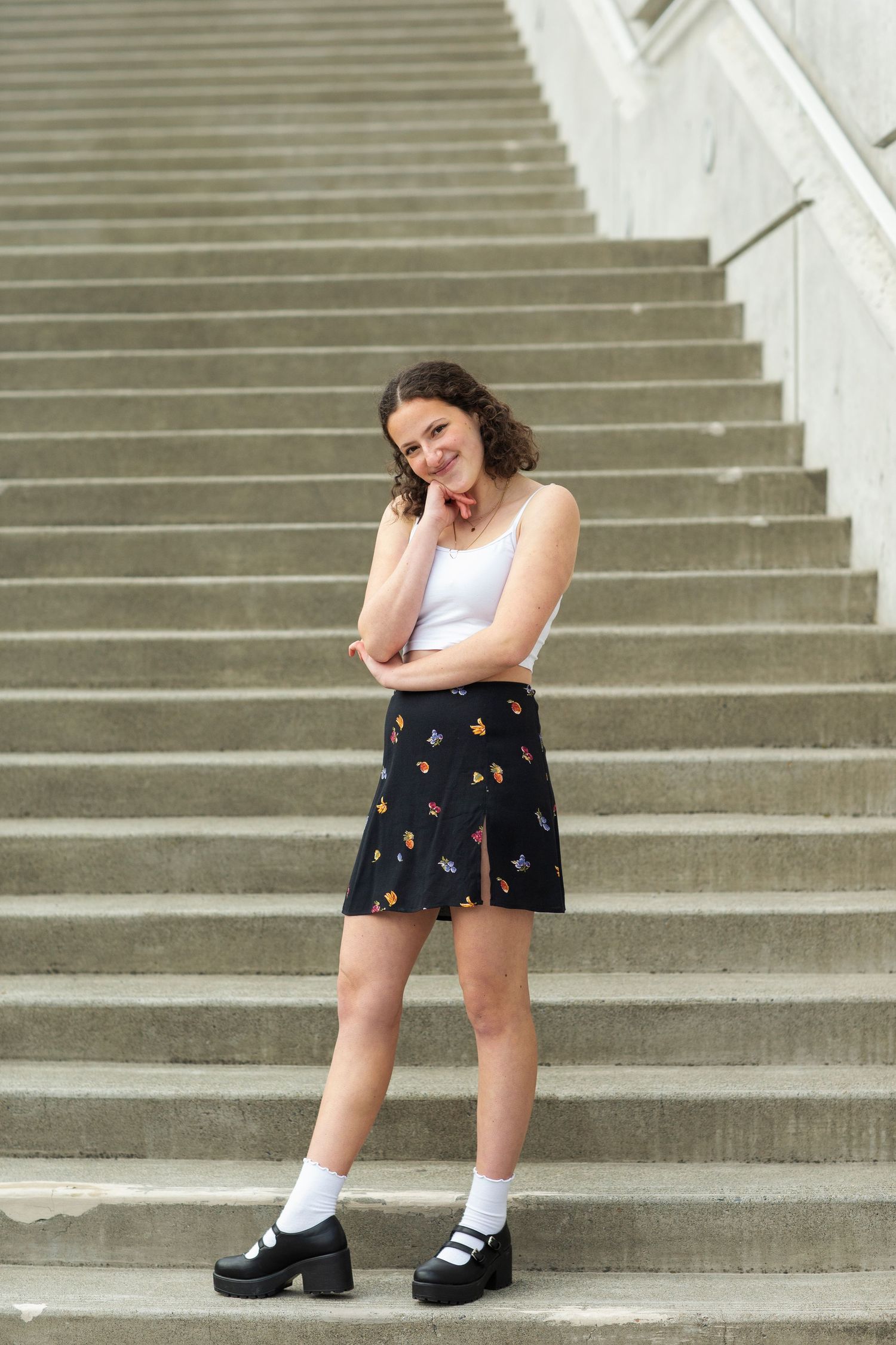 Young woman in white tank top and black floral skirt posing on outdoor concrete staircase.