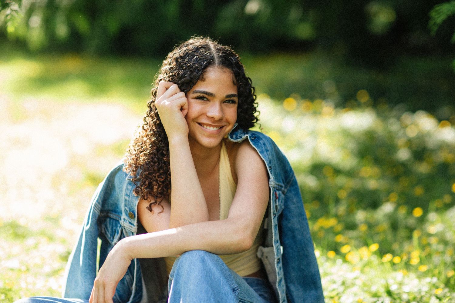 Young woman with curly hair smiling outdoors, wearing denim jacket and jeans, sitting on grass in sunny park.