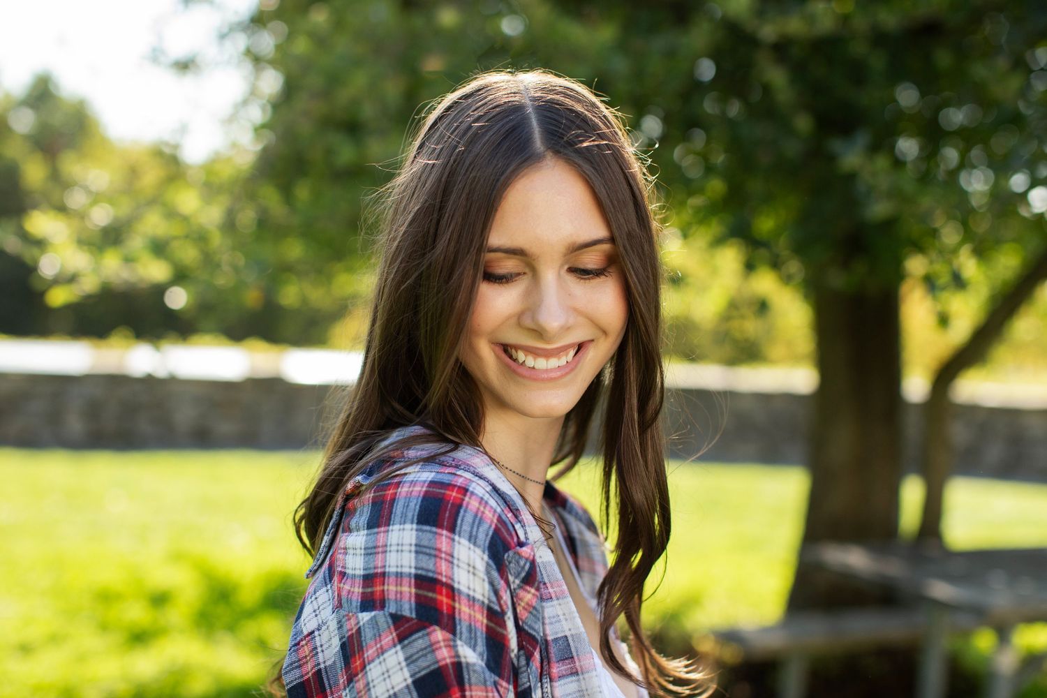 Young woman with long brown hair smiling downward wearing plaid shirt in sunny park.