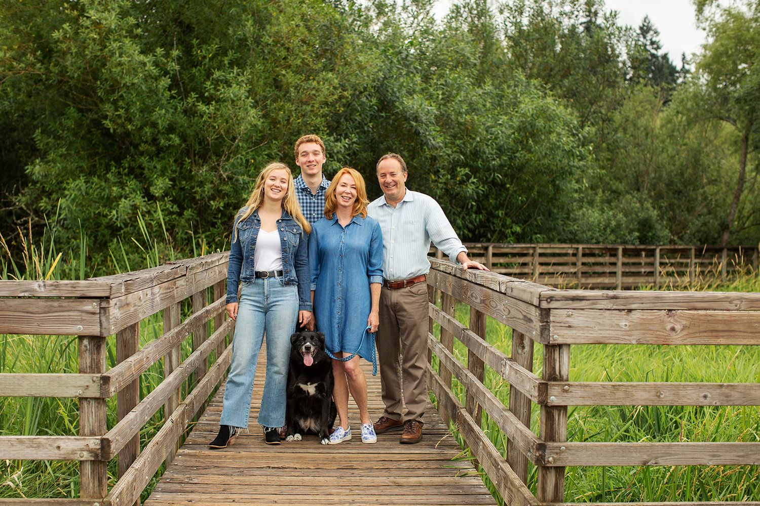 Family of four with a black dog posing on a wooden bridge surrounded by lush green trees outdoors.