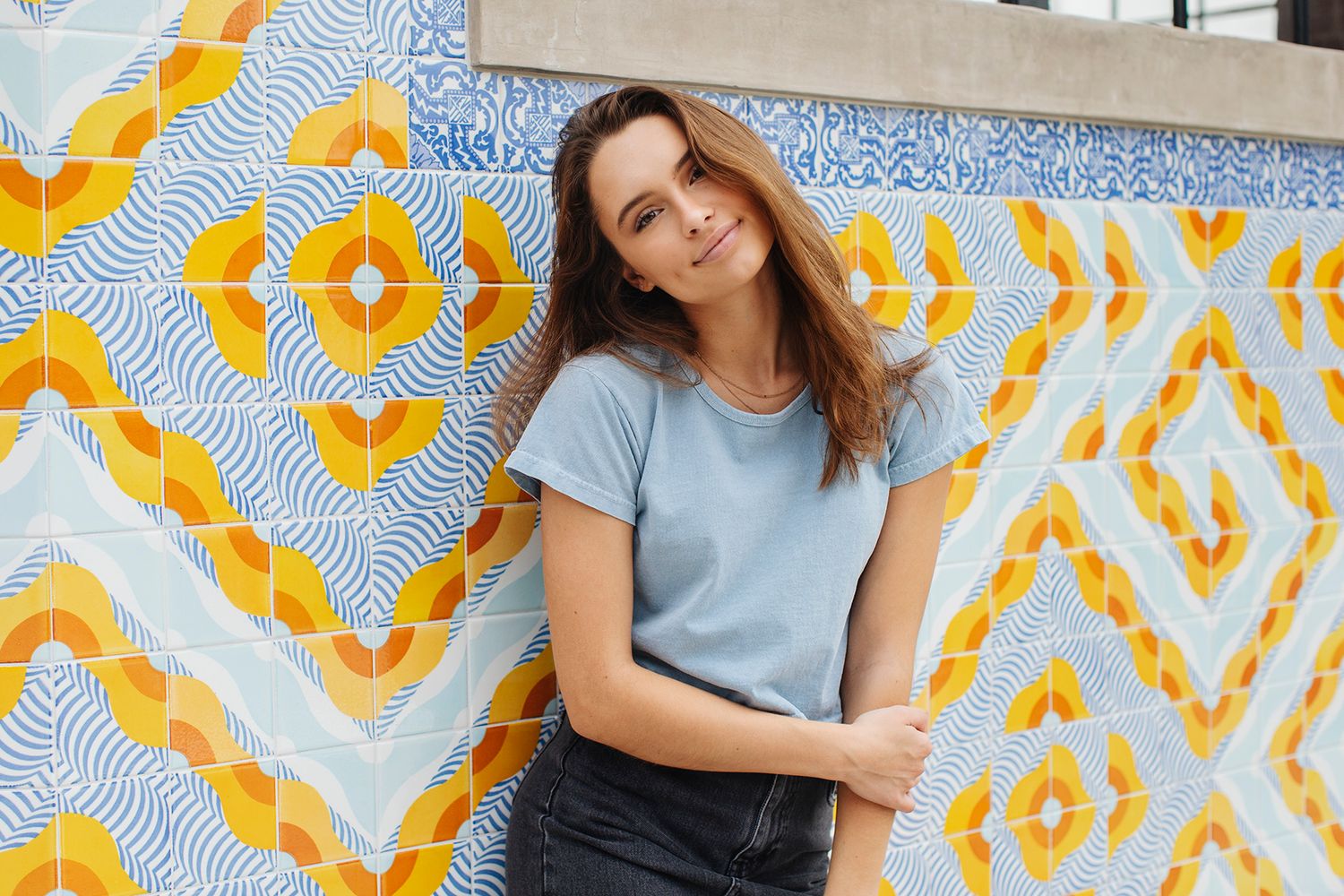 Young woman in light blue t-shirt posing against colorful geometric yellow and blue tiled wall.