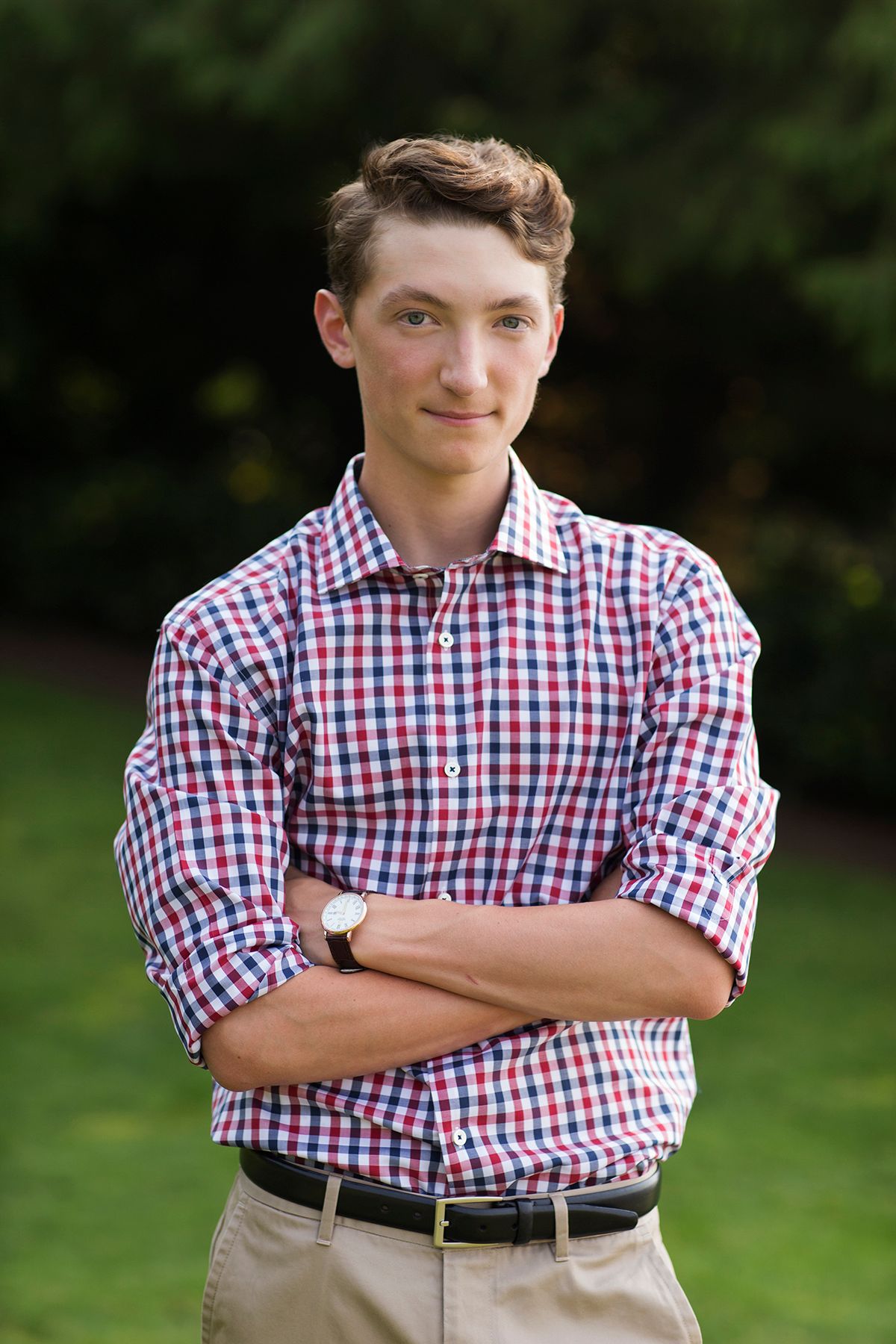 Young man in plaid shirt stands outdoors with arms crossed, smiling confidently against a green background.