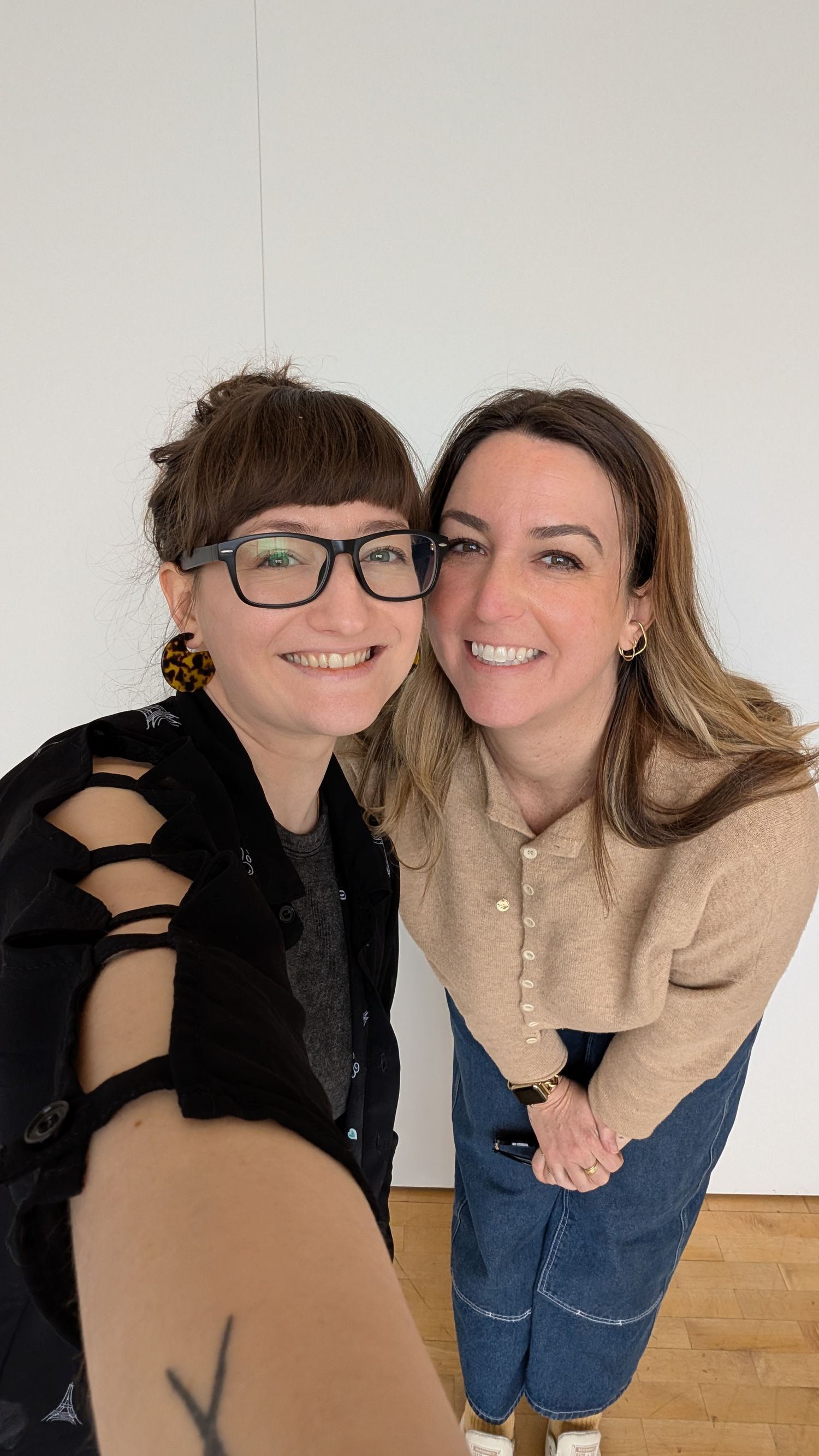 Two smiling women taking a selfie together in a bright white room, one wearing glasses and black outfit.