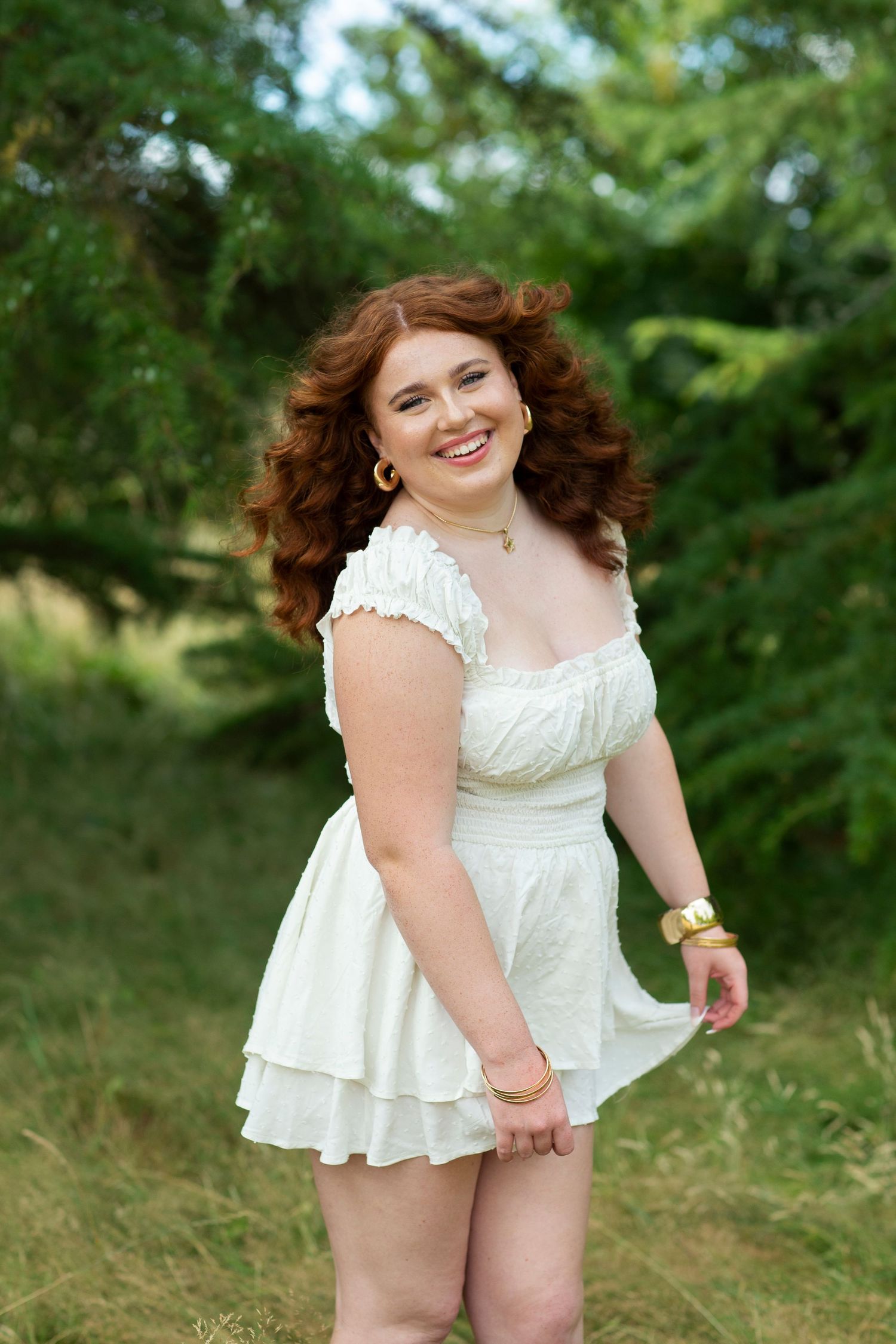 Smiling young woman with red curly hair in a white tiered mini dress posing in a green outdoor setting.