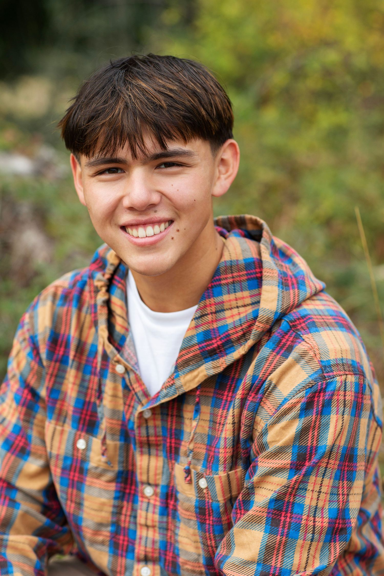 Smiling teenage boy in plaid hooded shirt seated outdoors in natural setting.