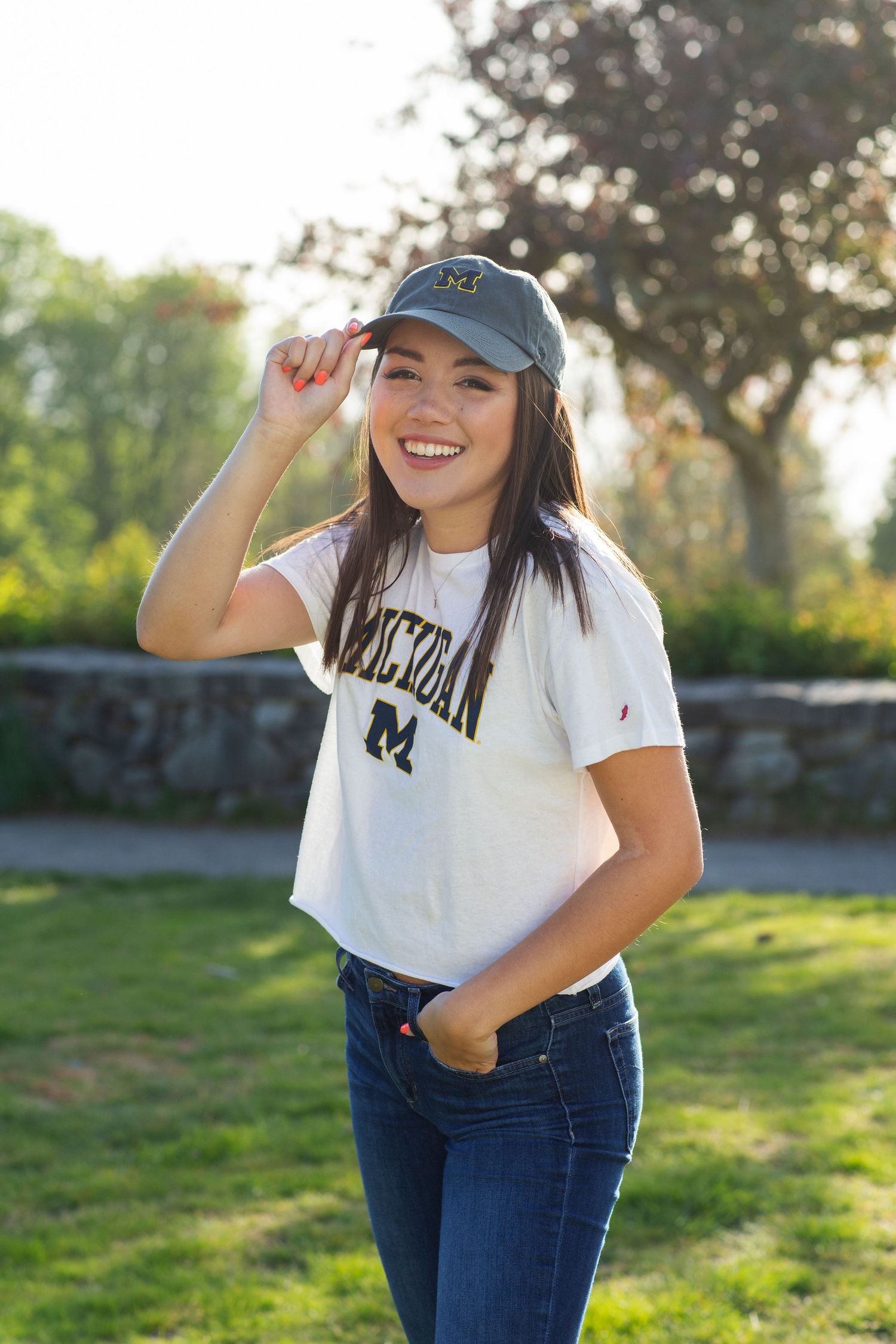 Smiling young woman in Michigan Wolverines cap and white tee poses outdoors in sunlit park setting.