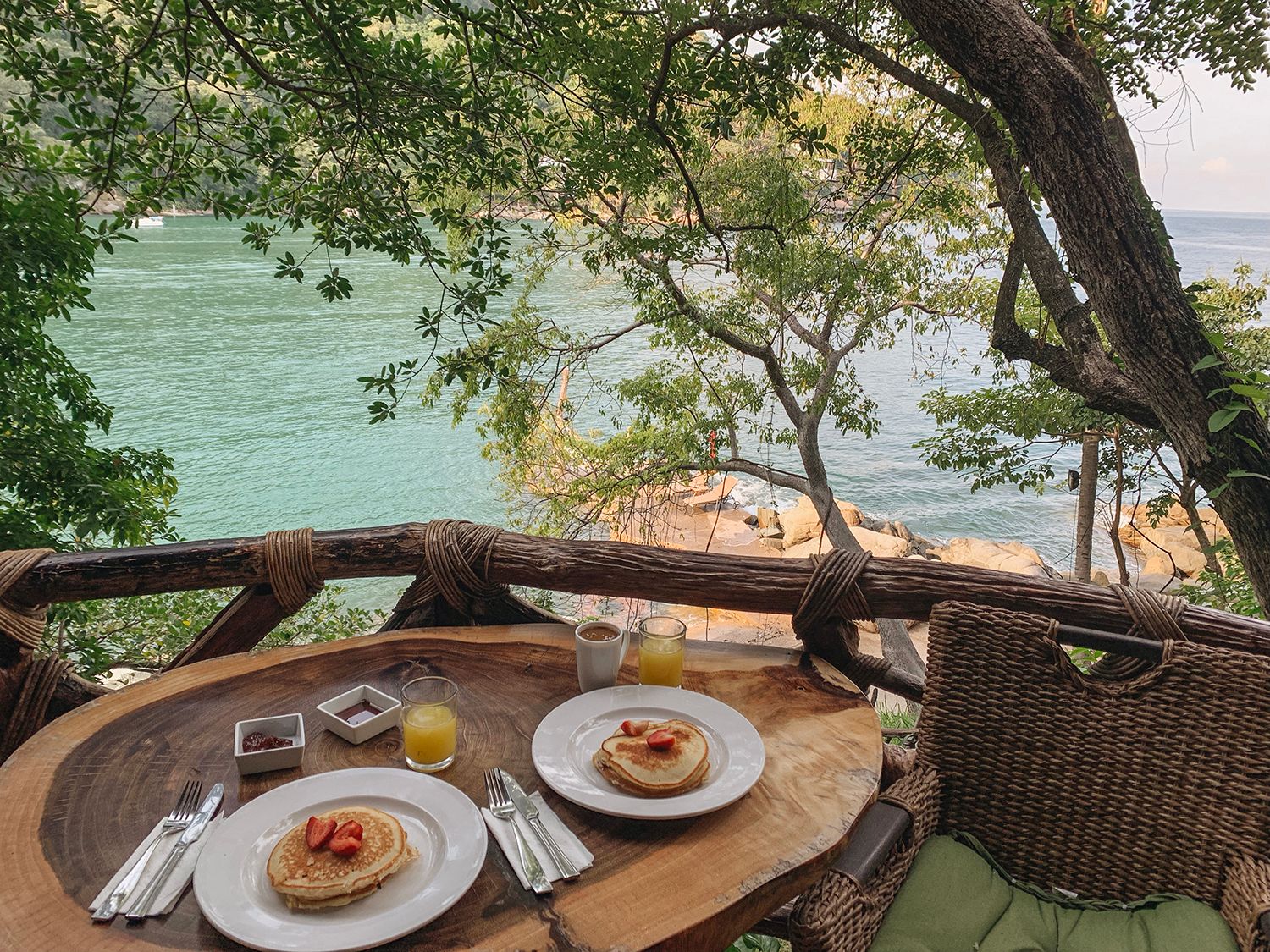 Breakfast for two with pancakes and orange juice on a wooden table overlooking a turquoise tropical bay.