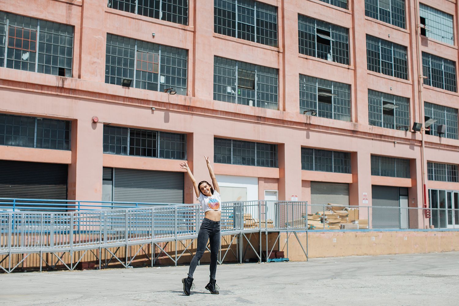 A woman dances joyfully outdoors with arms raised near a large red brick industrial building on a sunny day.