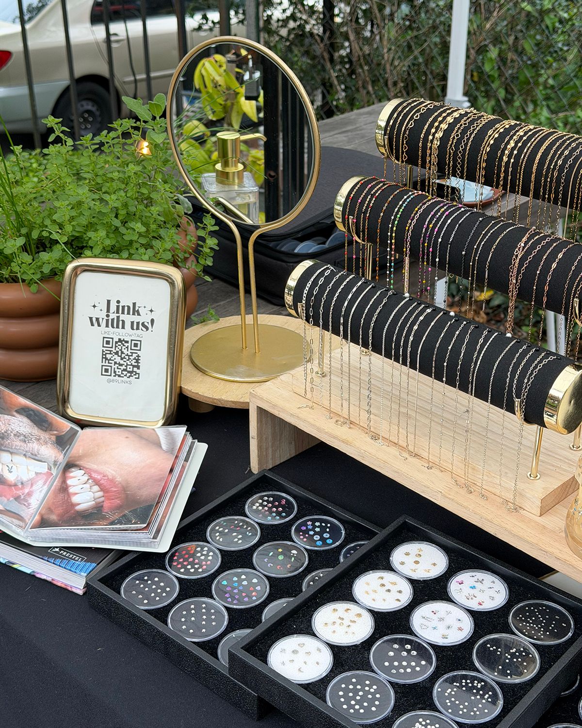 Outdoor jewelry vendor display with gold and black bracelet stands, circular earring trays, a mirror, and a QR code sign.