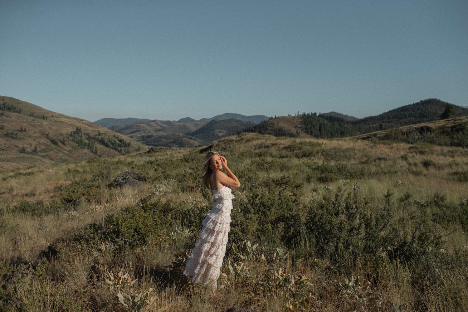 Blonde woman in a white lace dress standing in an open field with rolling green hills and blue sky.