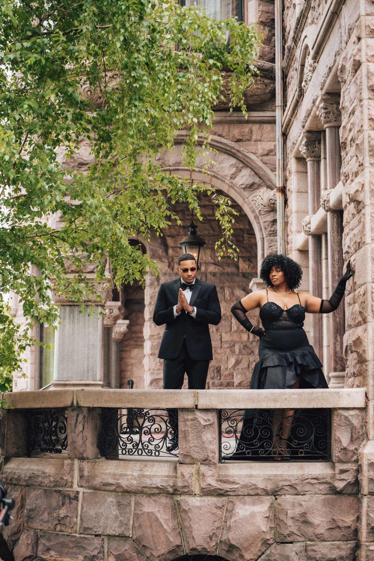 Elegant couple in formal black attire posing on a stone balcony of a historic building, man in tuxedo, woman in black gown.