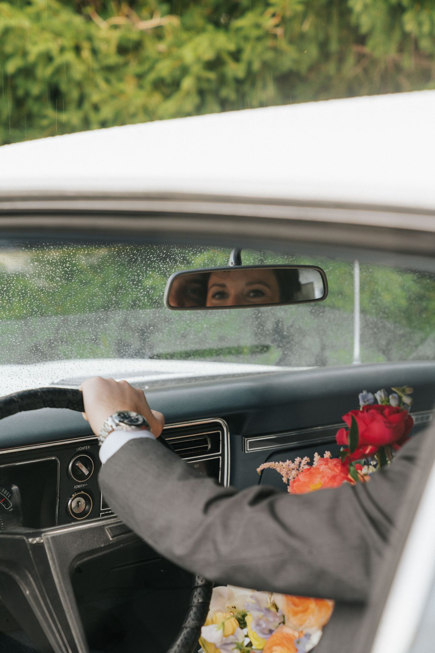 Groom in suit with floral boutonniere driving vintage car, bride's eyes reflected in rearview mirror.