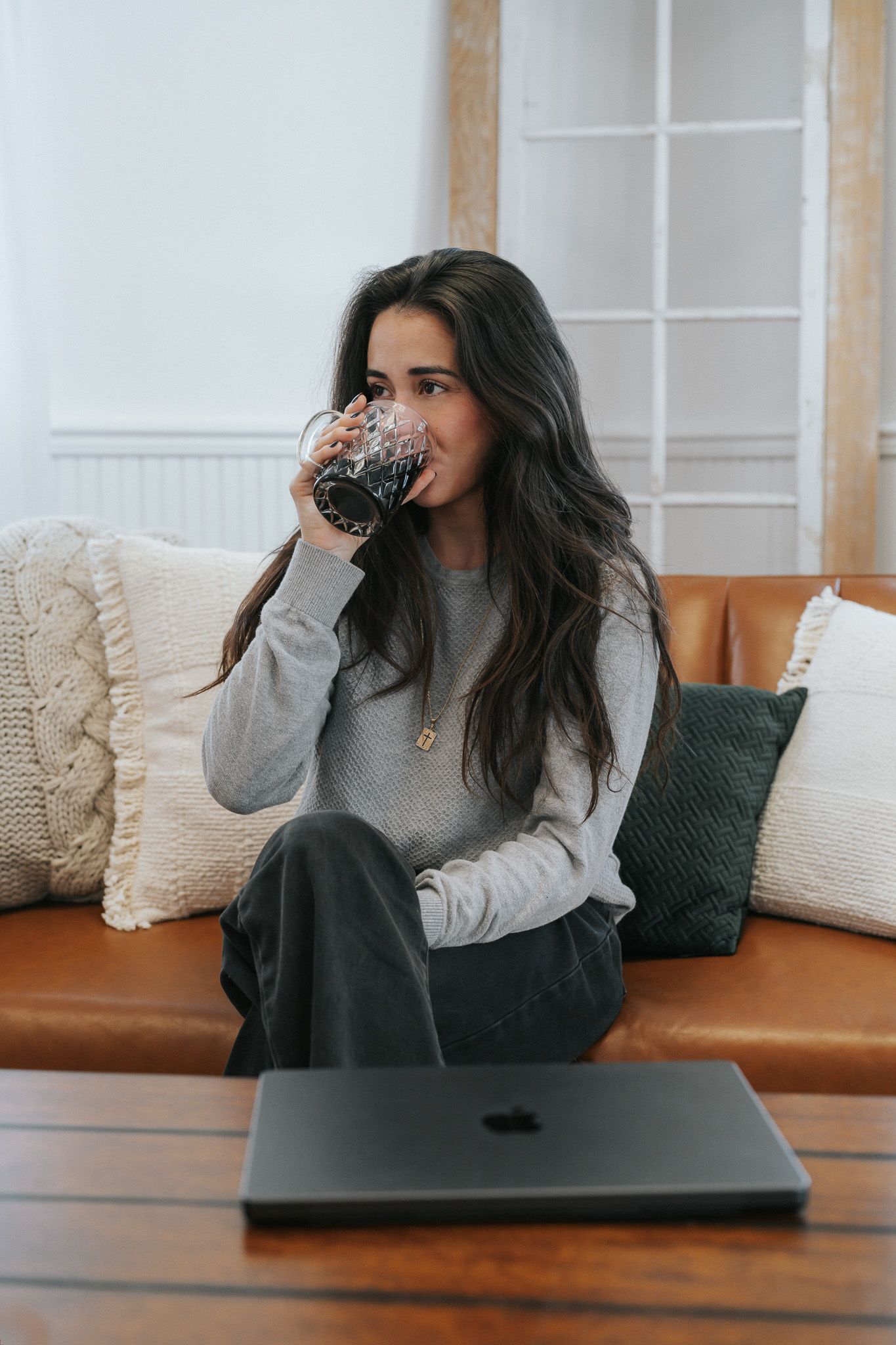 Young woman in gray sweater drinking from glass while sitting on leather sofa with laptop.