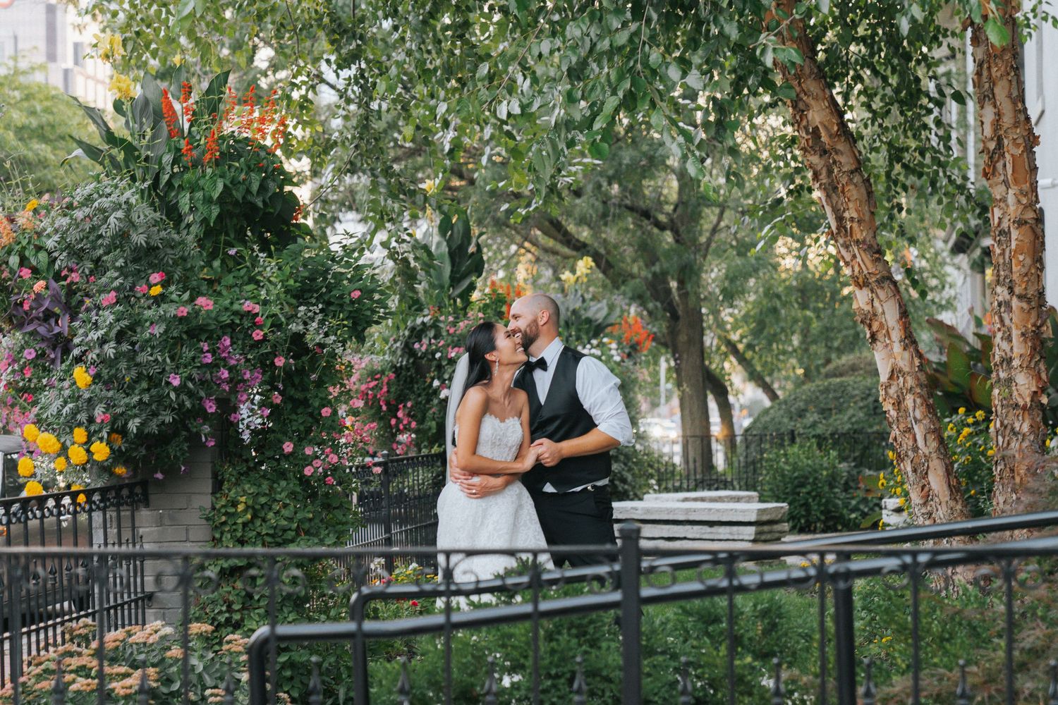 Bride and groom share a romantic kiss surrounded by lush greenery and colorful flowers in an outdoor garden setting.