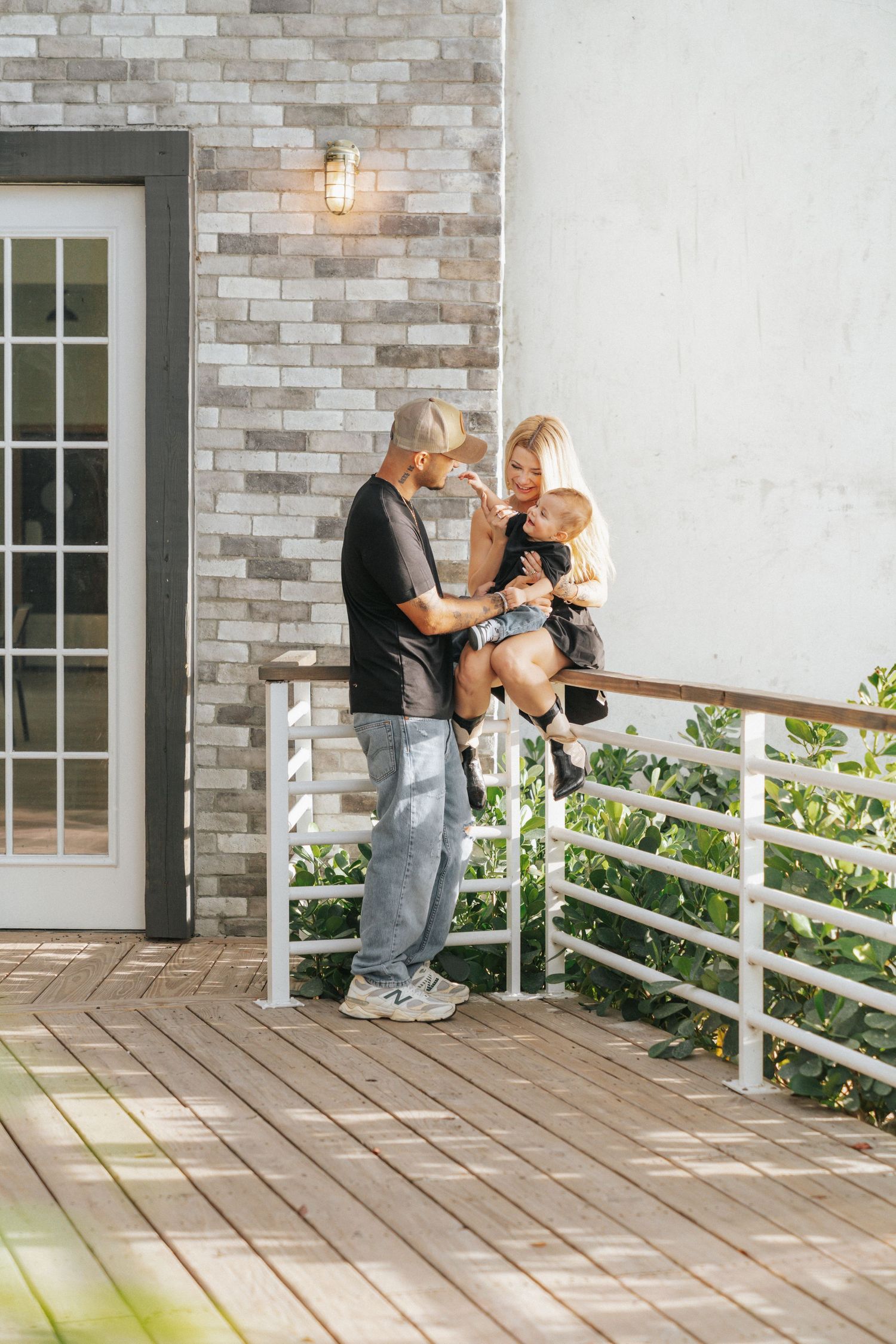 Happy family with parents holding a baby on a wooden porch with white railings outside a modern brick home.