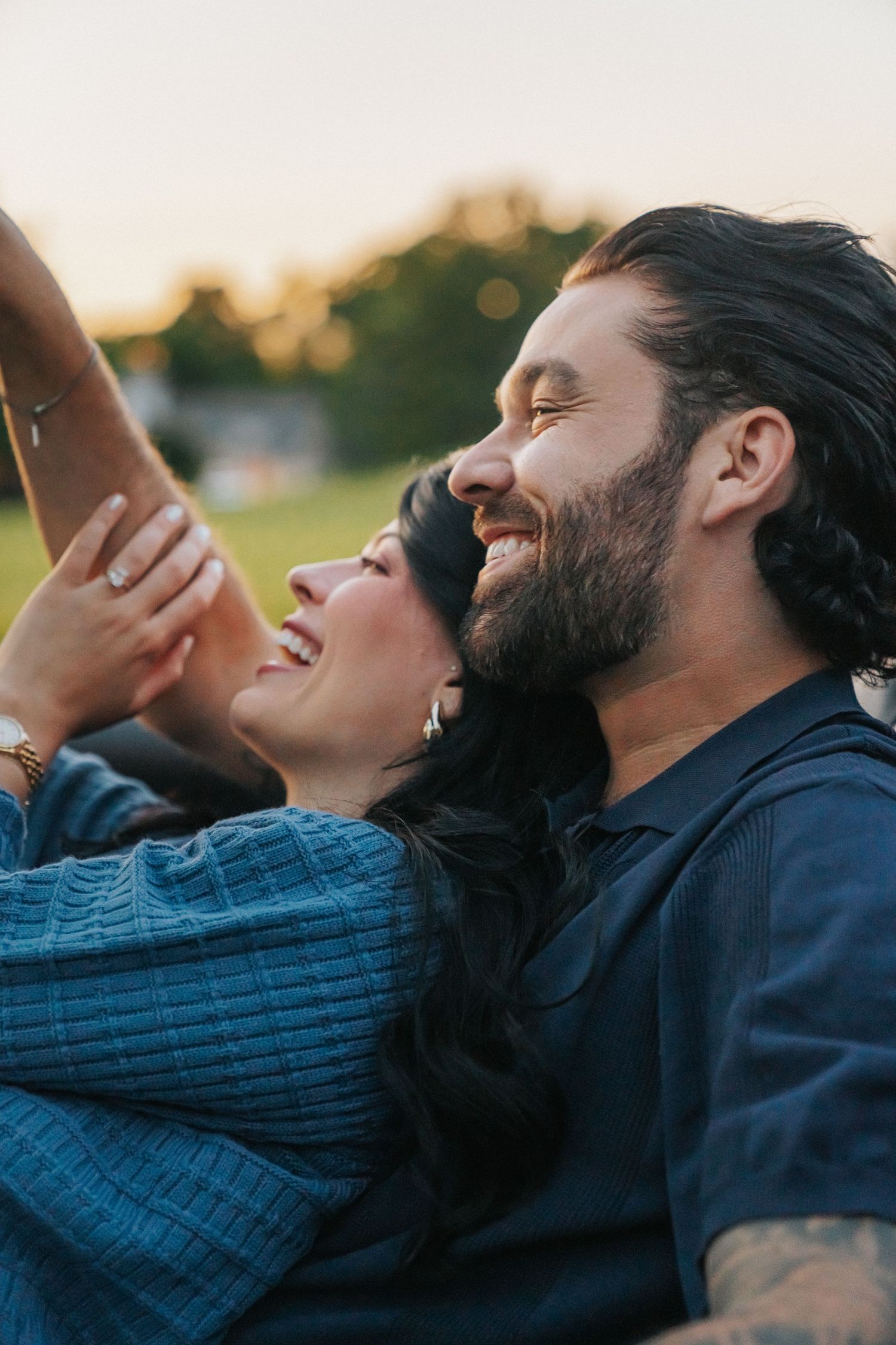 A happy couple laughs together outdoors at sunset, woman leaning on bearded man's shoulder.