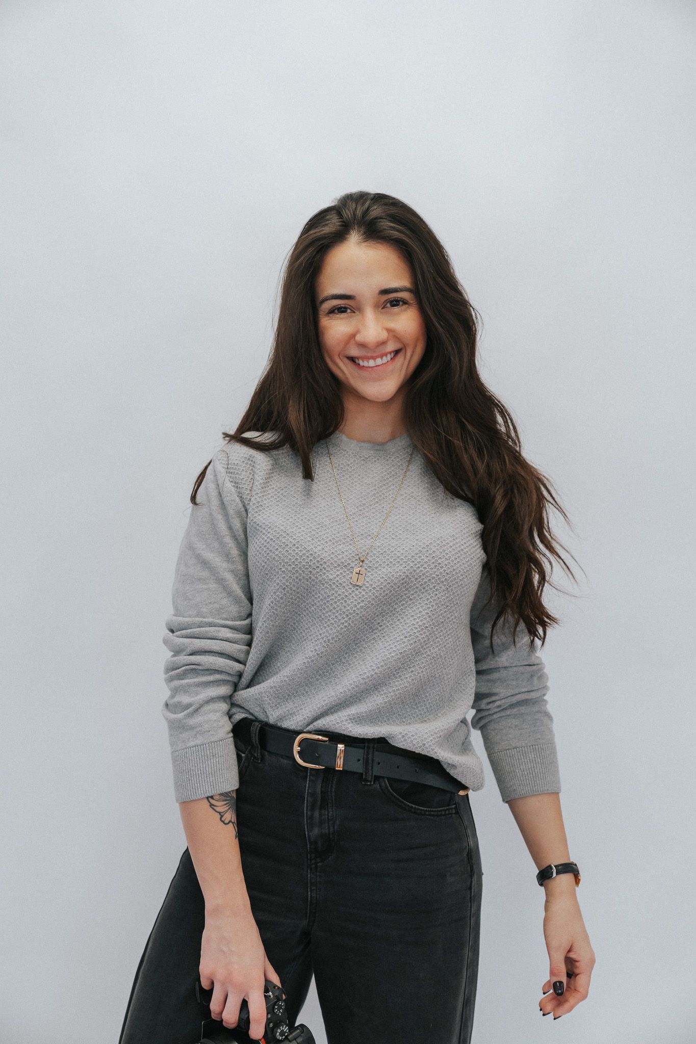 Smiling brunette woman in a gray sweater and black jeans posing against a white background.