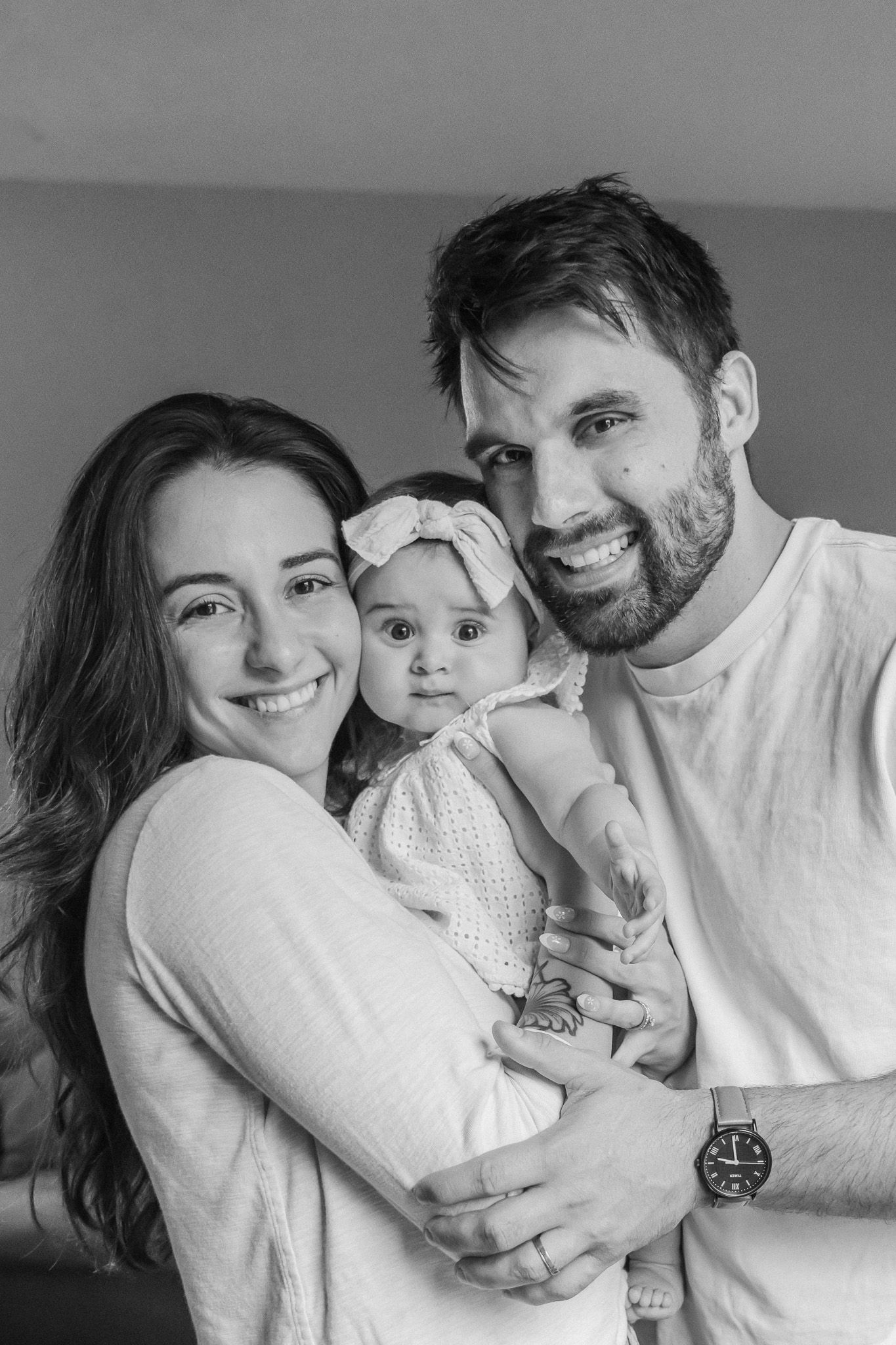 Black and white portrait of smiling young parents holding their baby girl with a bow headband indoors.