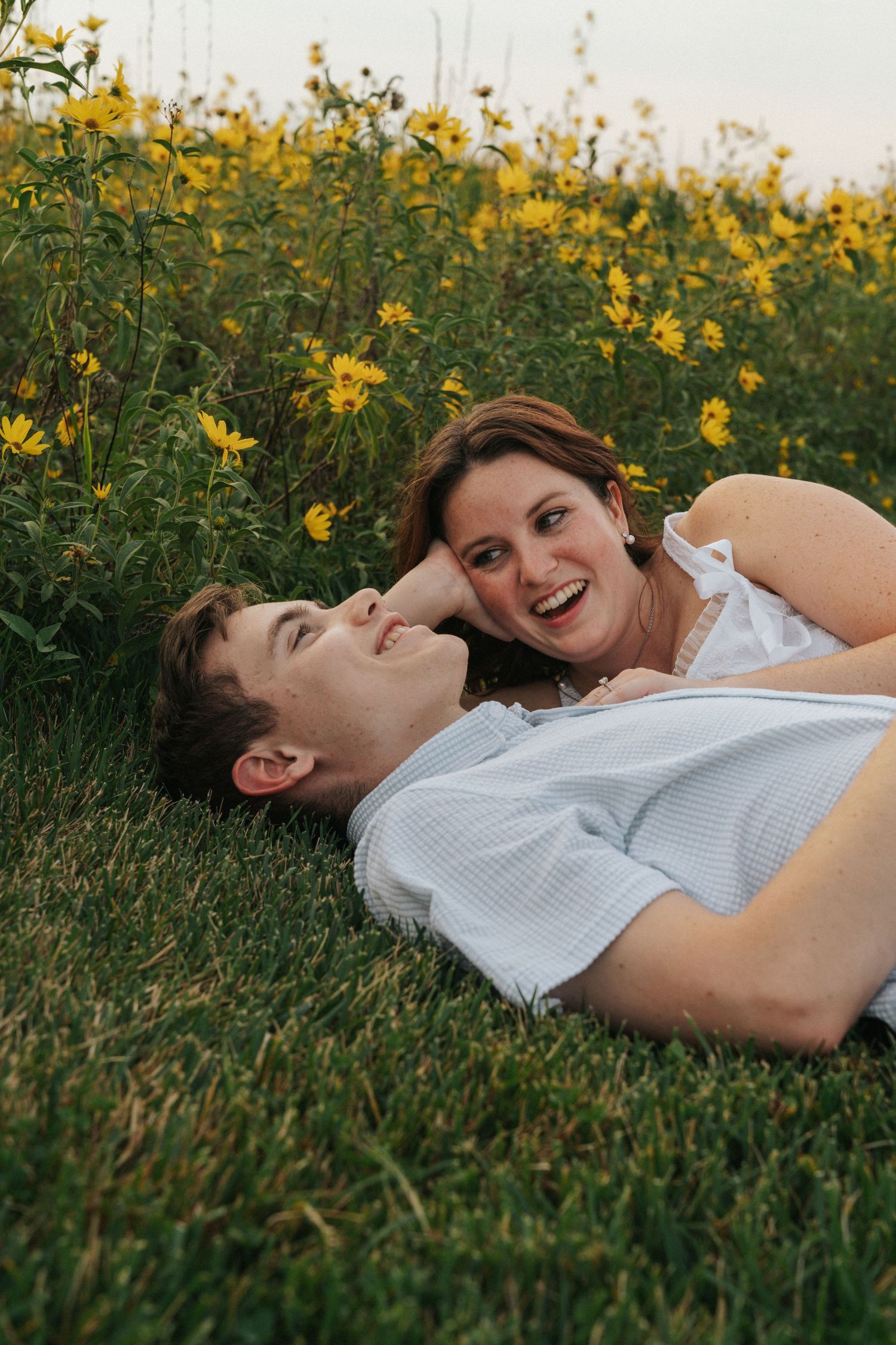 A joyful couple laughs together while lying in a field of bright yellow wildflowers at sunset.