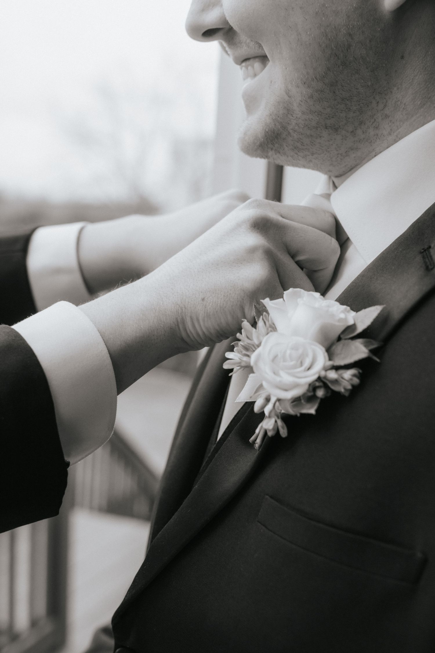 Black and white close-up of groom smiling as hands adjust his bow tie and floral boutonniere.