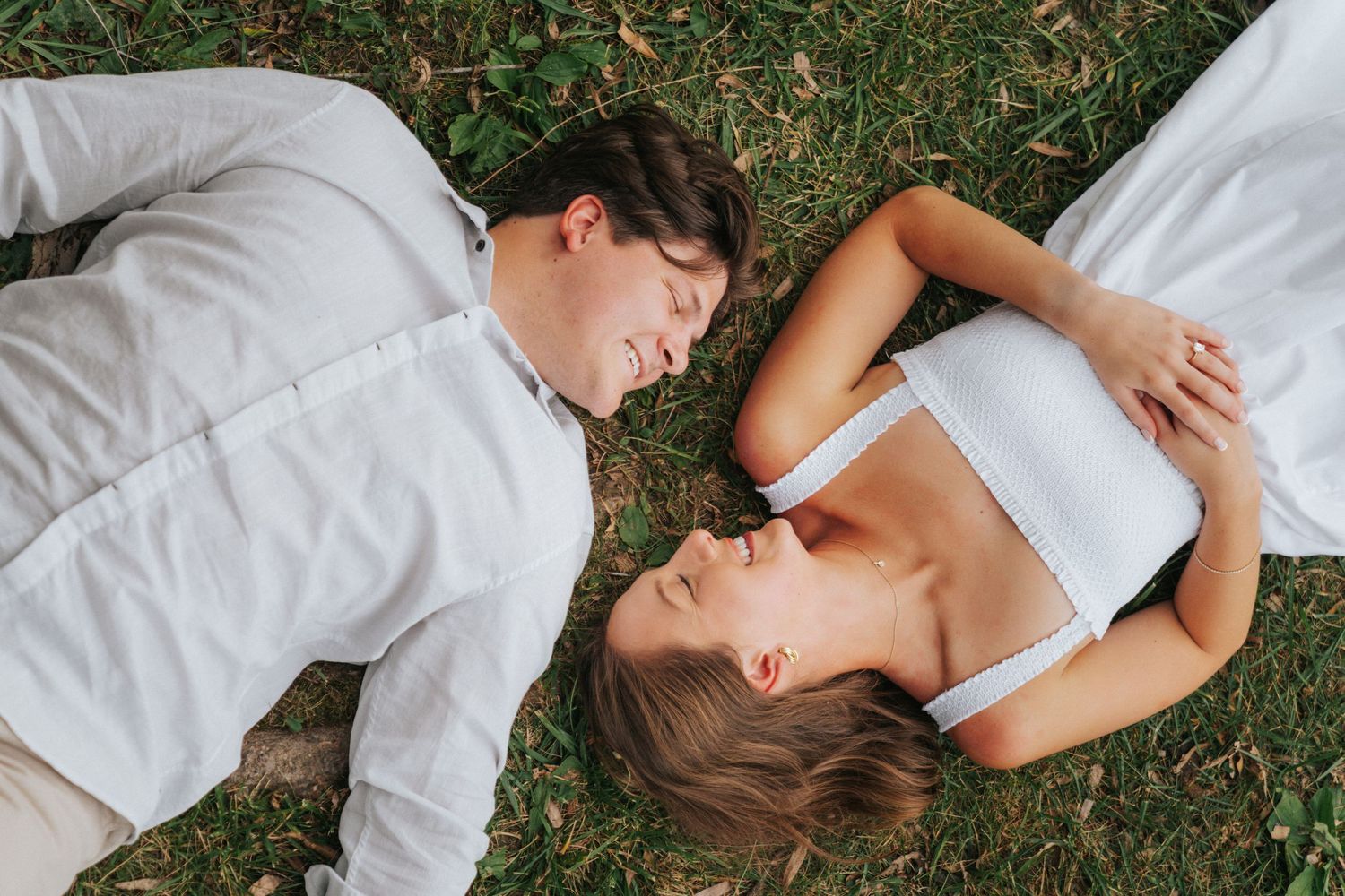 Romantic couple lying on green grass face to face, wearing white outfits, sharing an intimate moment outdoors.