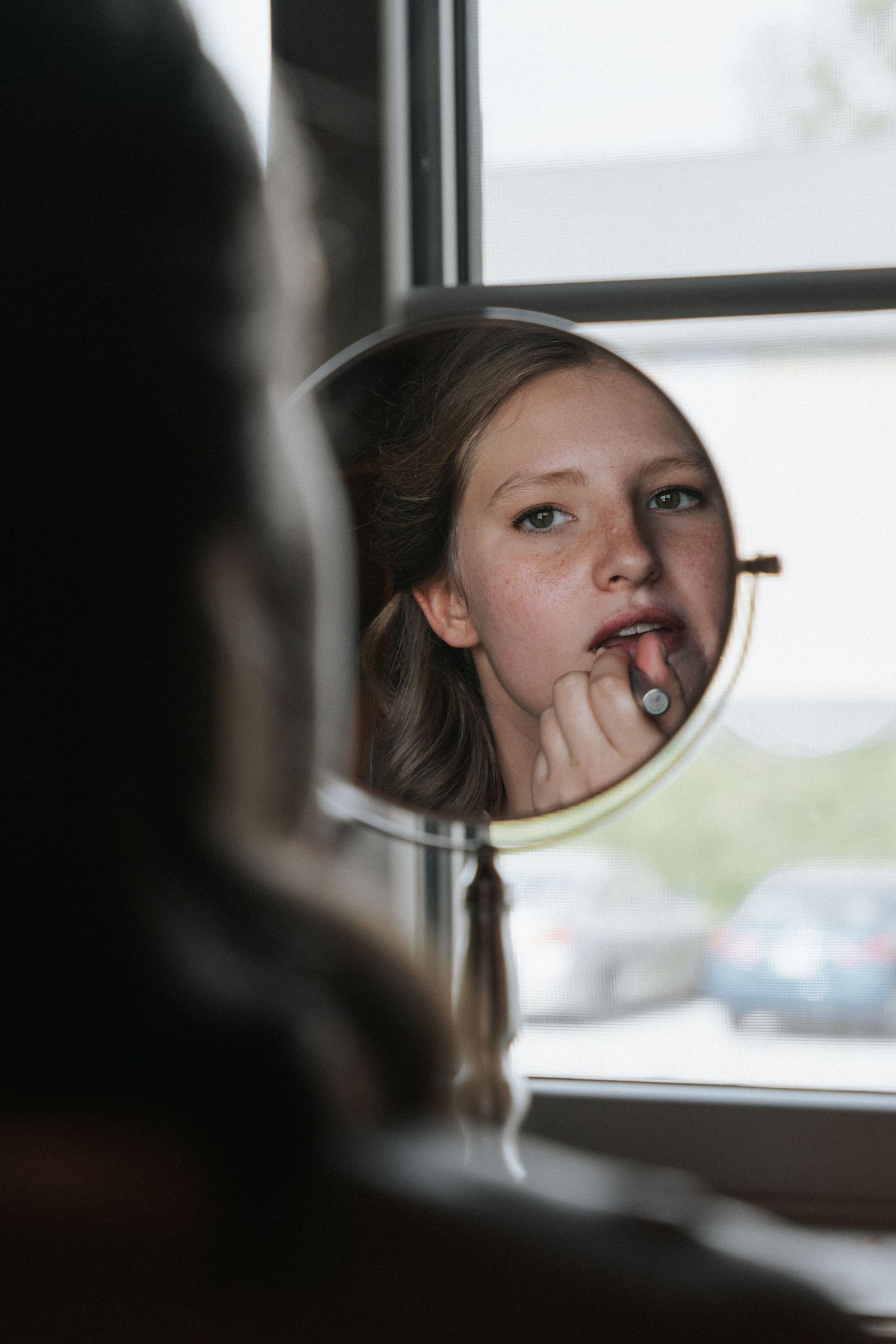 Young bridesmaid applying lipstick while looking at her reflection in a round mirror.