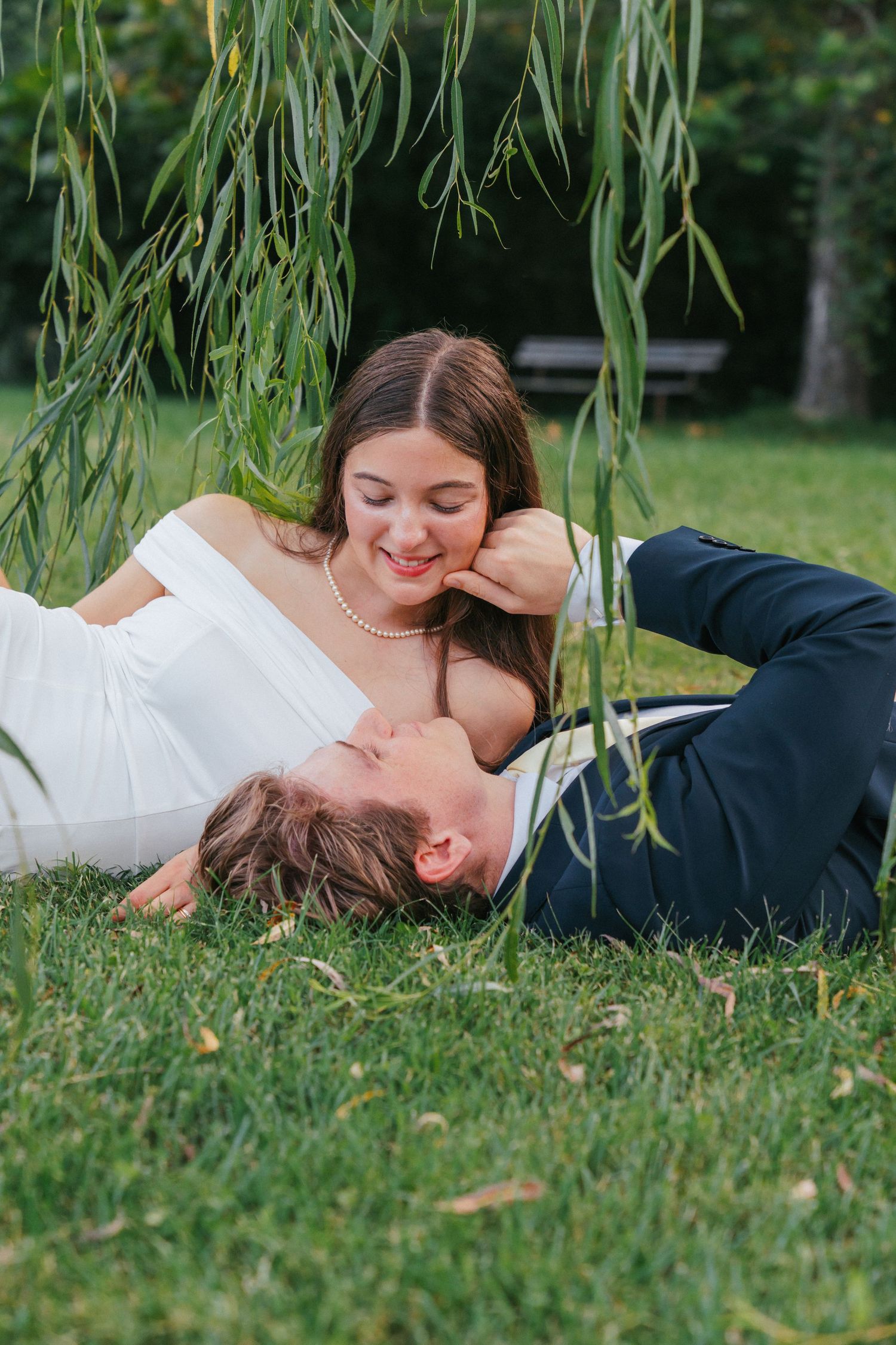Bride and groom lying on grass under willow tree, sharing a romantic moment in wedding attire outdoors.