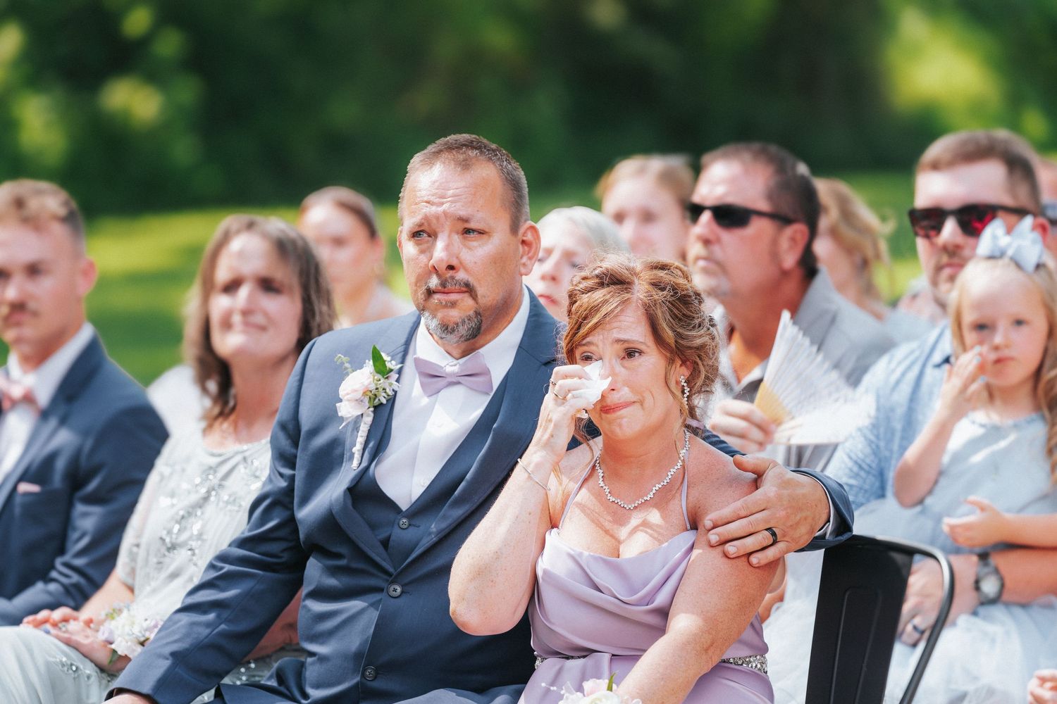 Emotional wedding guests wiping tears during outdoor ceremony, man in gray suit comforting woman in lavender dress.