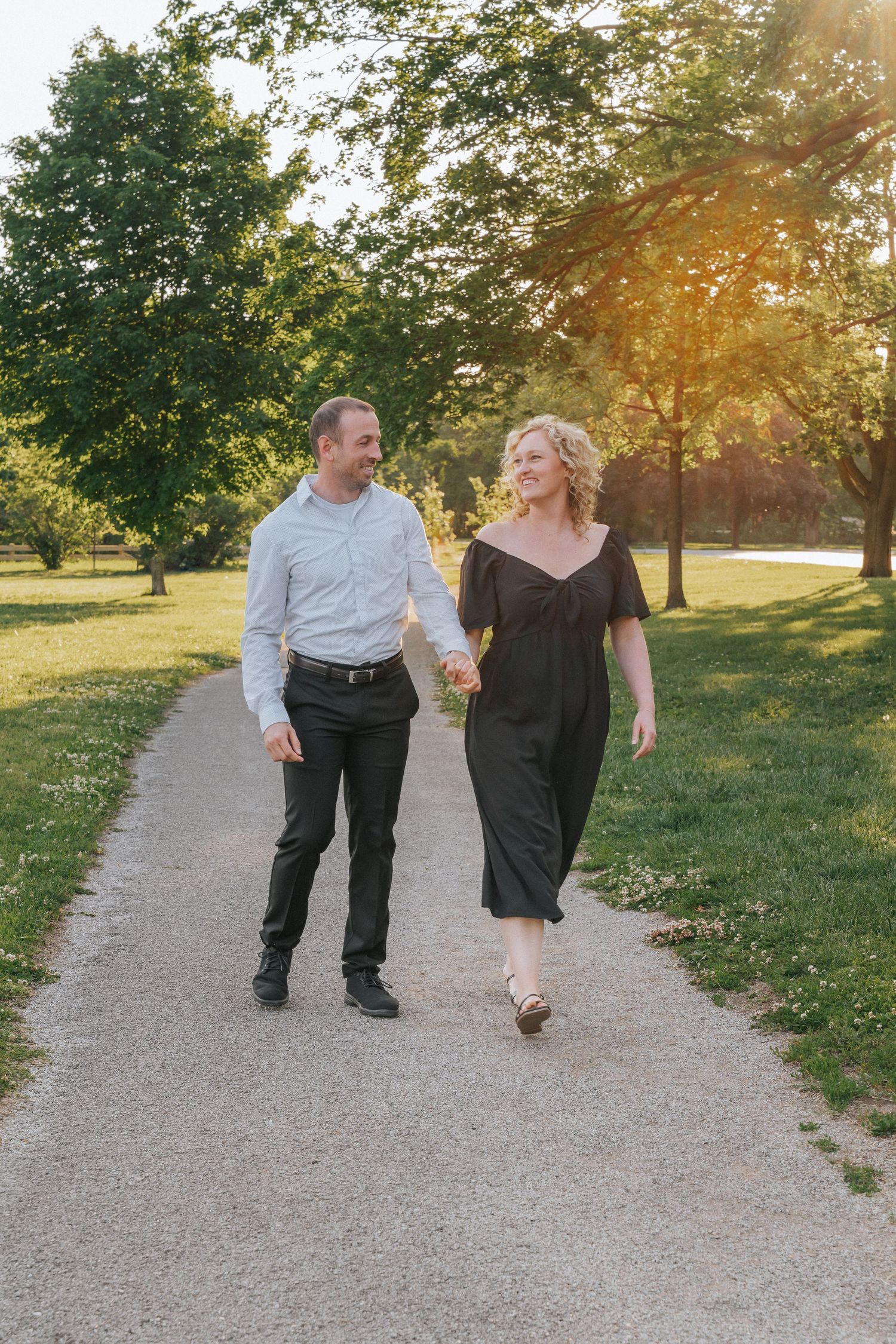 A couple holds hands walking on a park path at golden hour, man in white shirt, woman in black dress.