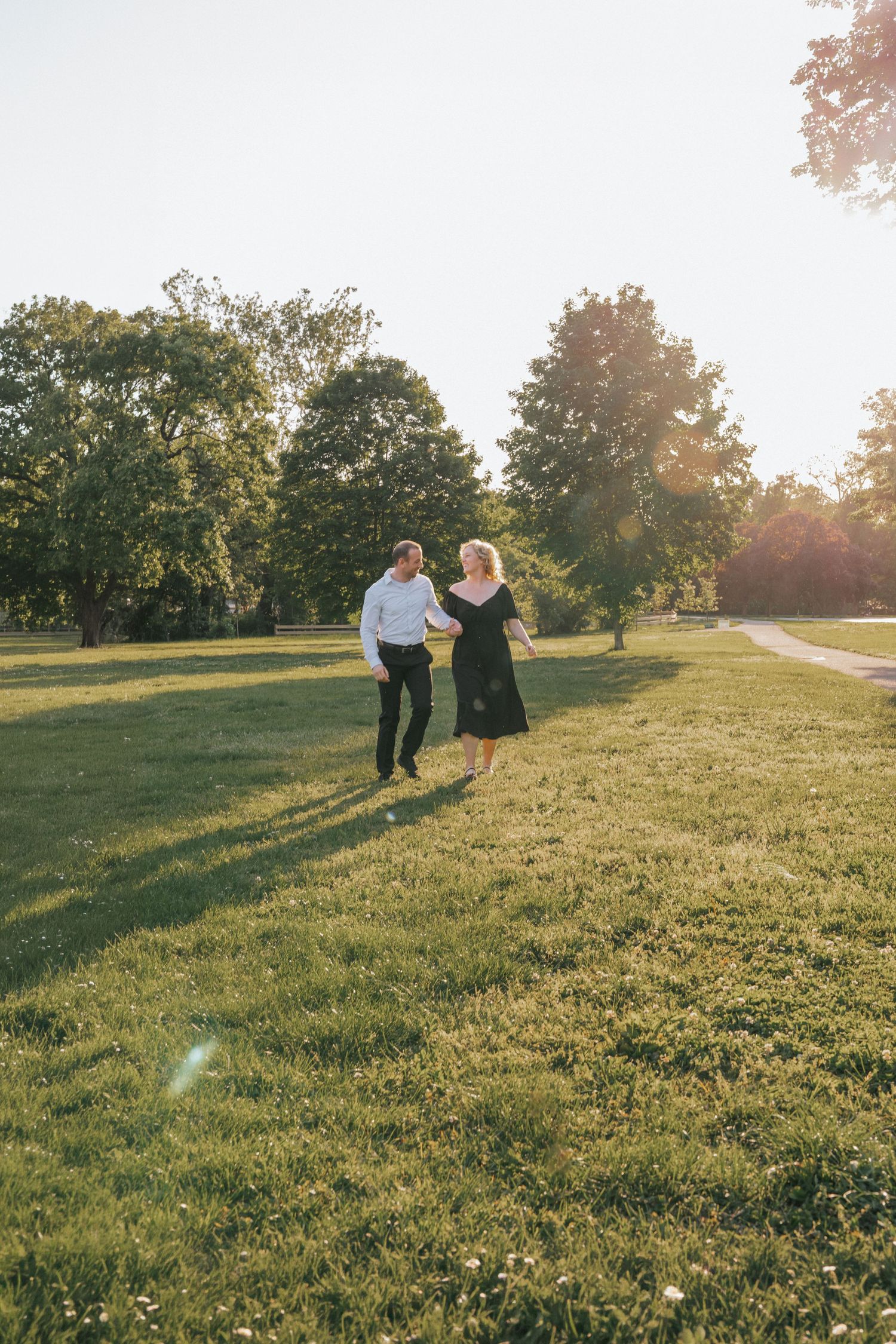 A couple holds hands and runs through a sunny green park, surrounded by lush trees in warm golden light.