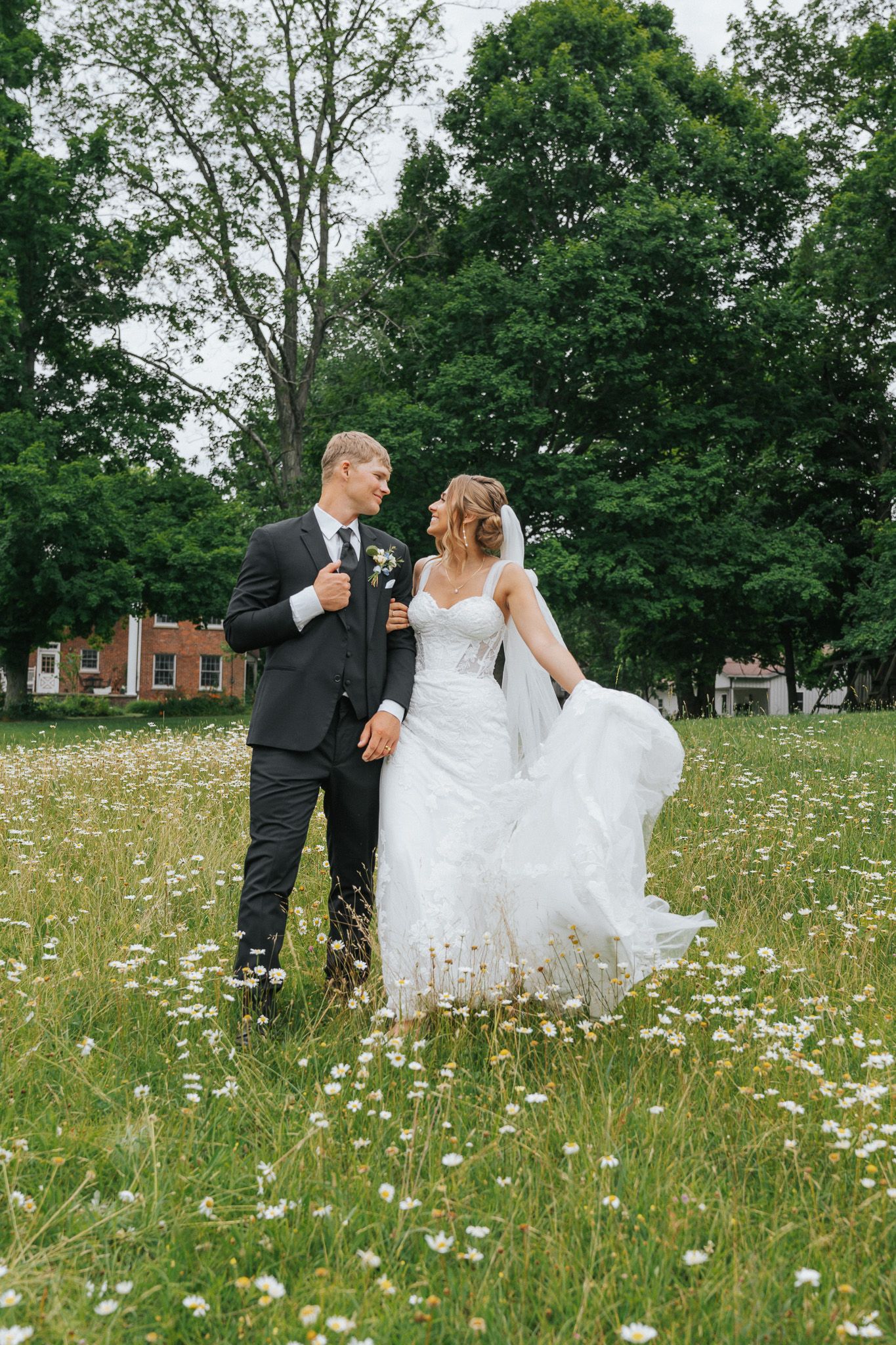 Bride and groom standing in a daisy field with lush green trees, bride's white gown flowing in the breeze.