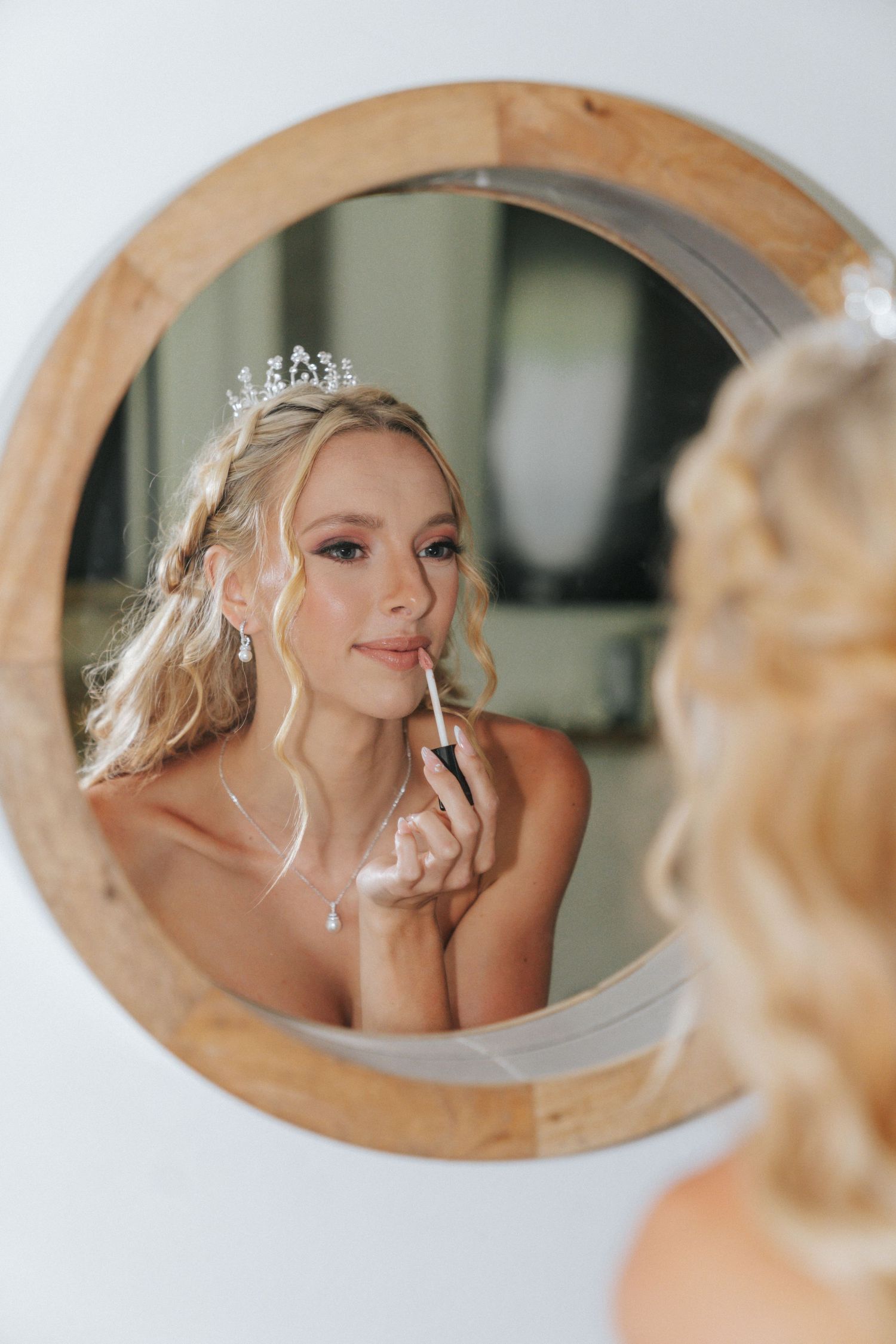 Bride applying lipstick while looking in a round wooden mirror, wearing a tiara and elegant jewelry.