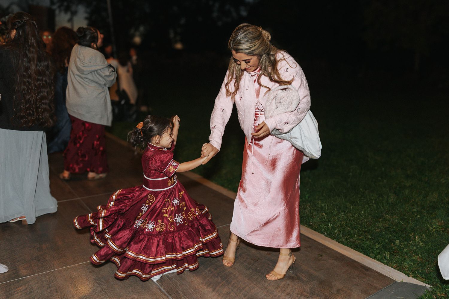Woman in pink dress dancing with young girl in red floral outfit on outdoor dance floor at night.