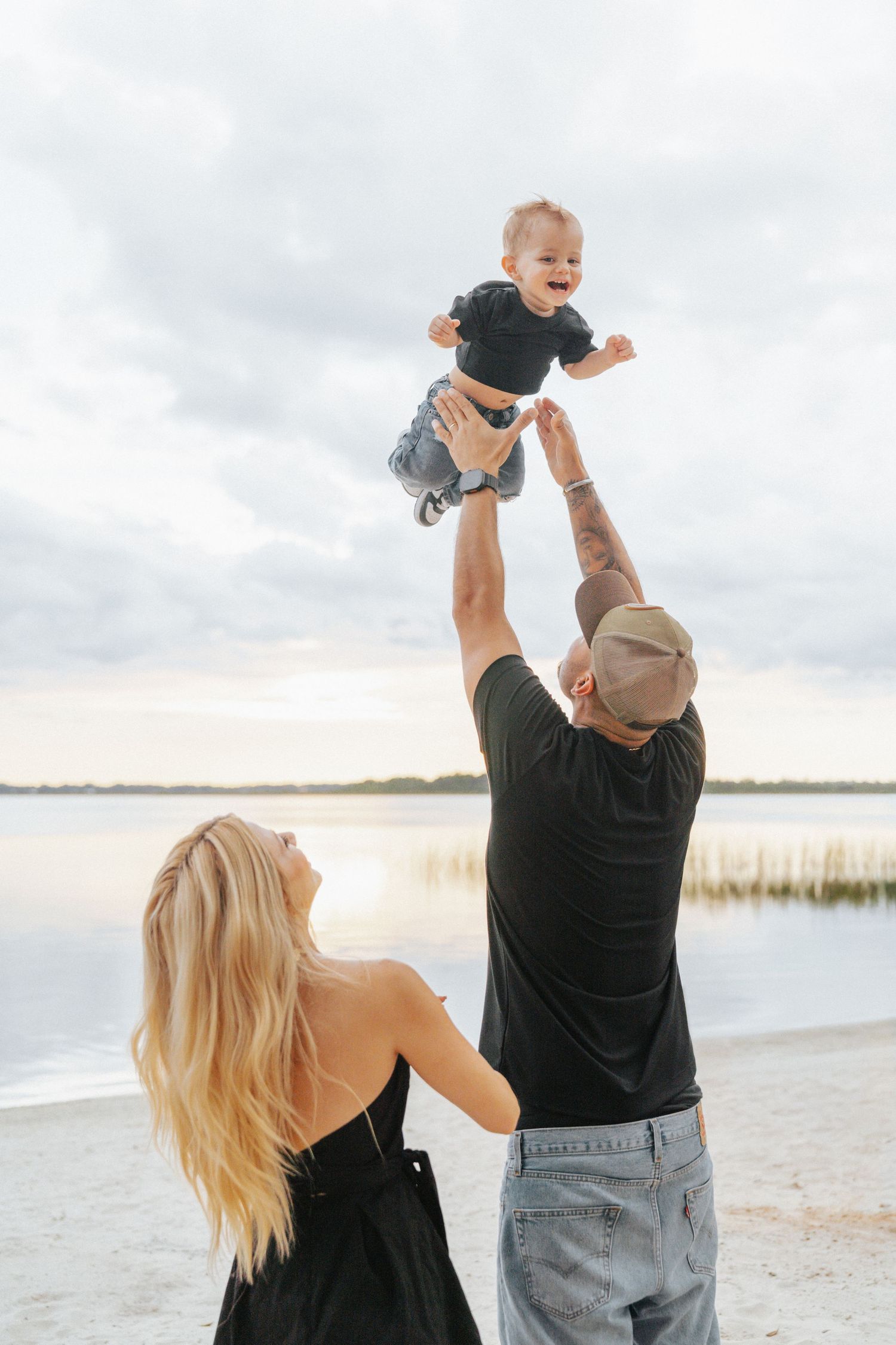 A happy family at the beach, with a tattooed father lifting a laughing baby in the air while the blonde mother watches.