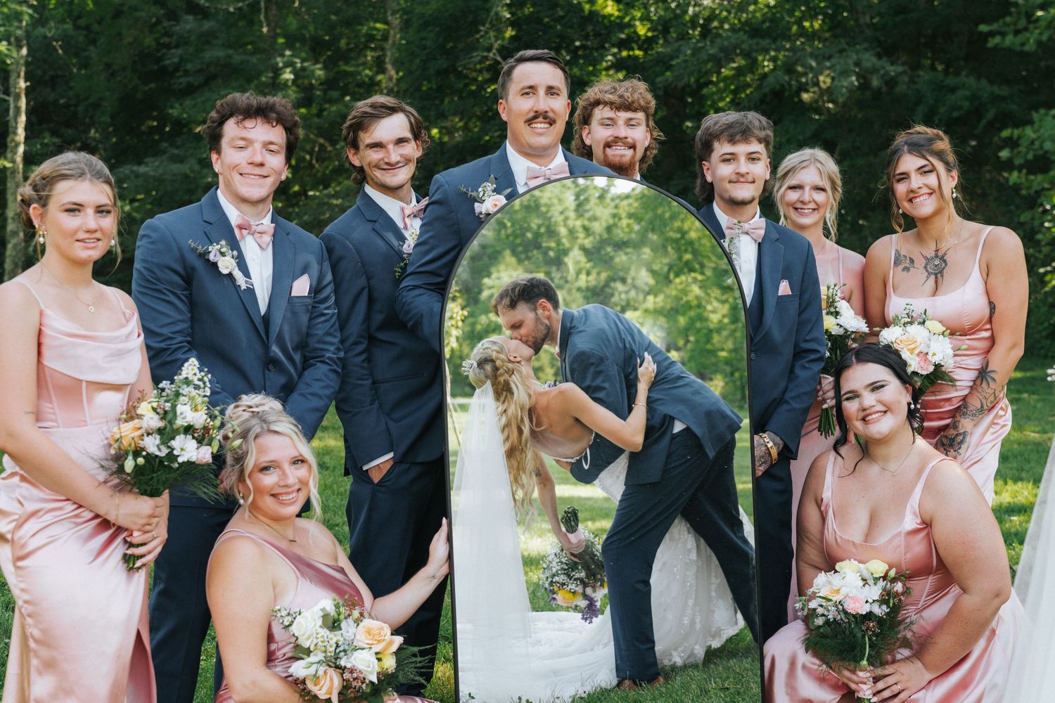 Wedding party poses outdoors with bride and groom kissing reflected in a mirror, bridesmaids in pink and groomsmen in navy.