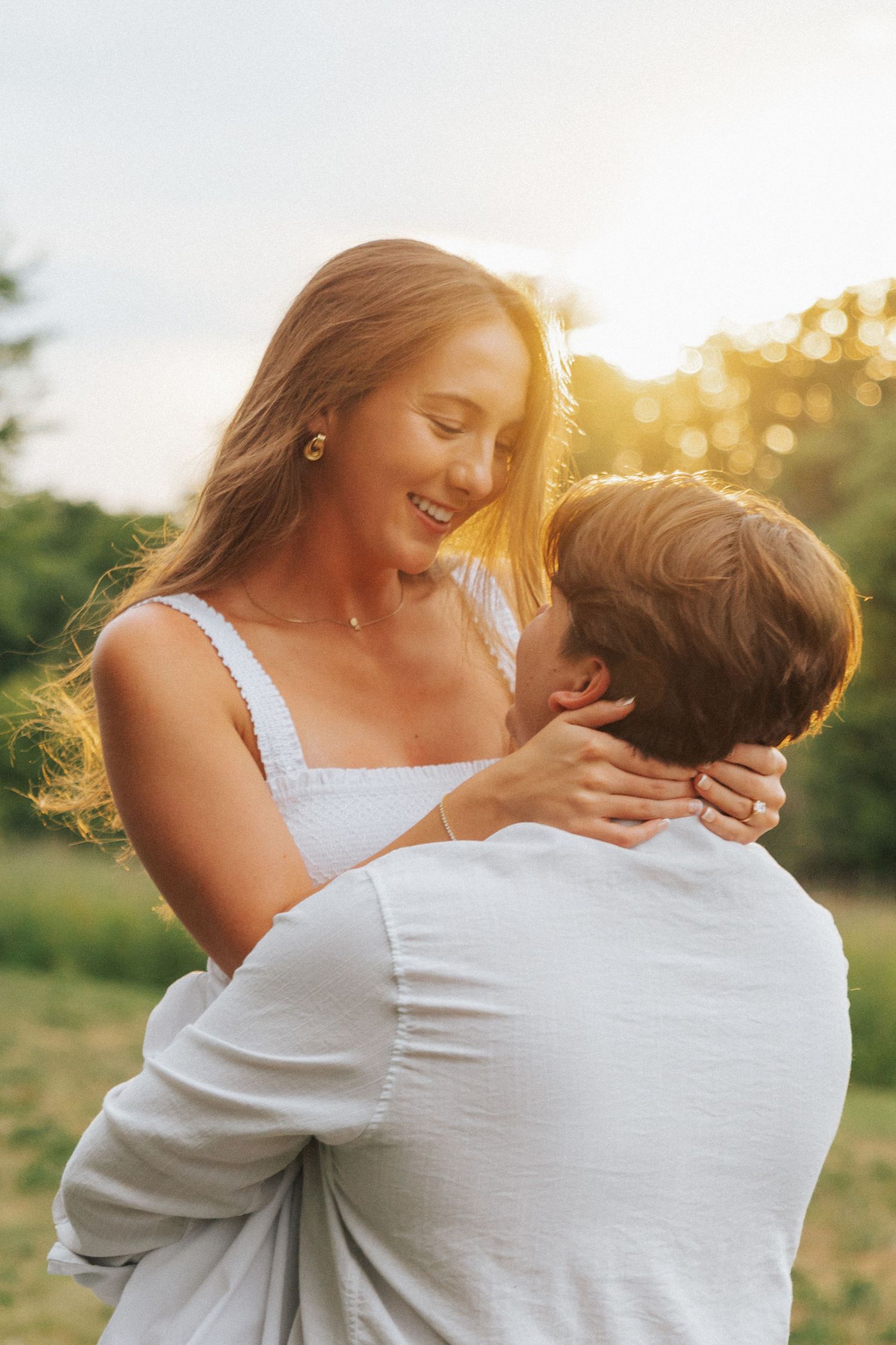 Couple embracing at golden hour sunset, woman smiling joyfully as man lifts her in a romantic outdoor setting.