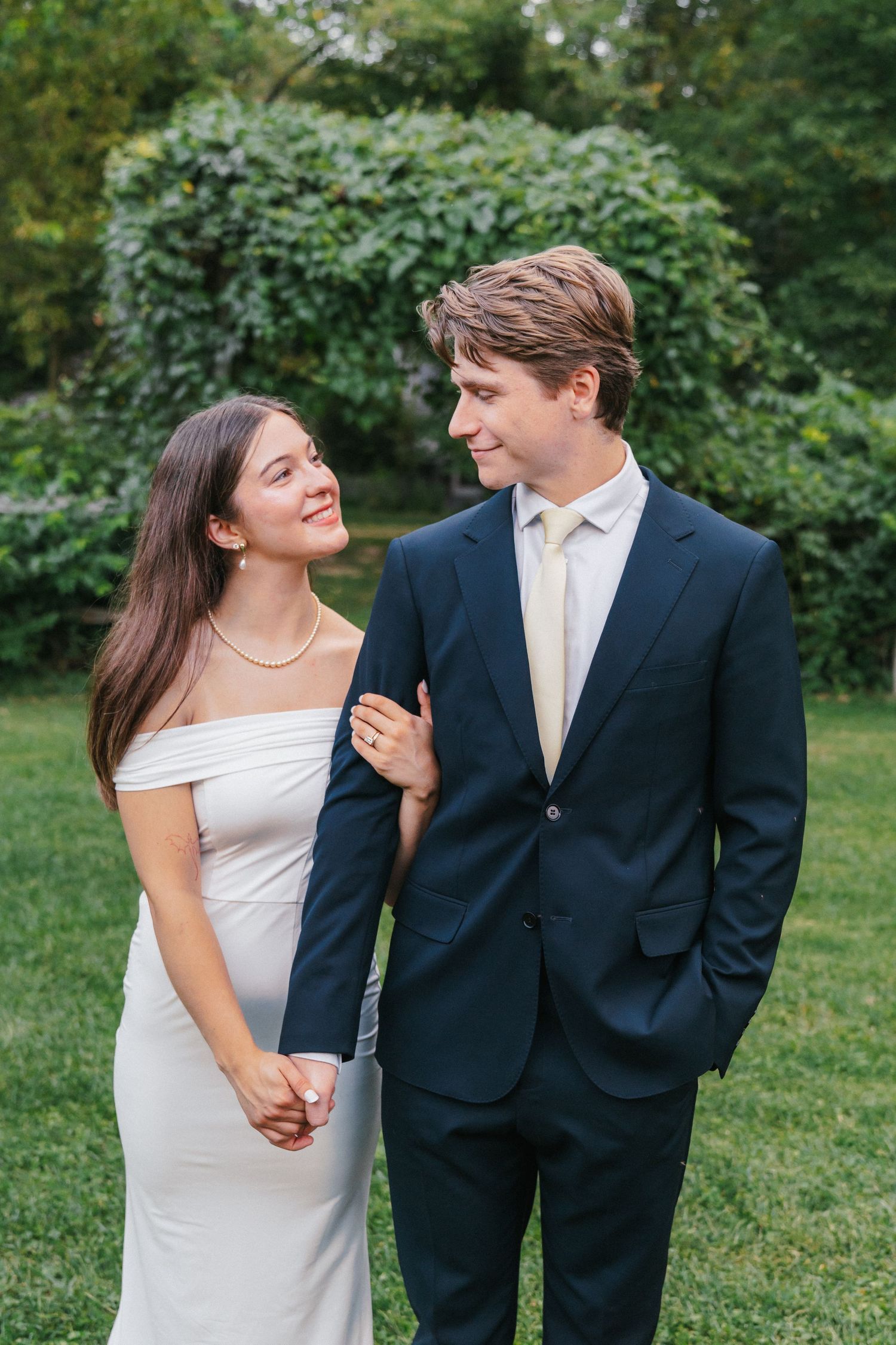 A couple in formal attire smiling at each other outdoors, woman in white gown and man in navy suit.