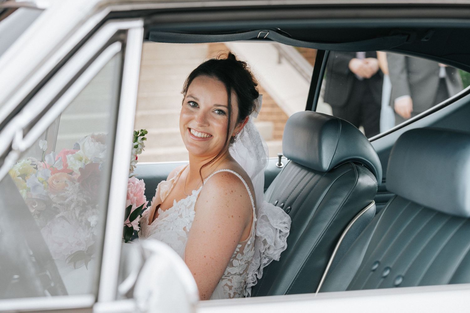 Smiling bride in a white lace dress sitting in a vintage car, holding a bouquet, on her wedding day.
