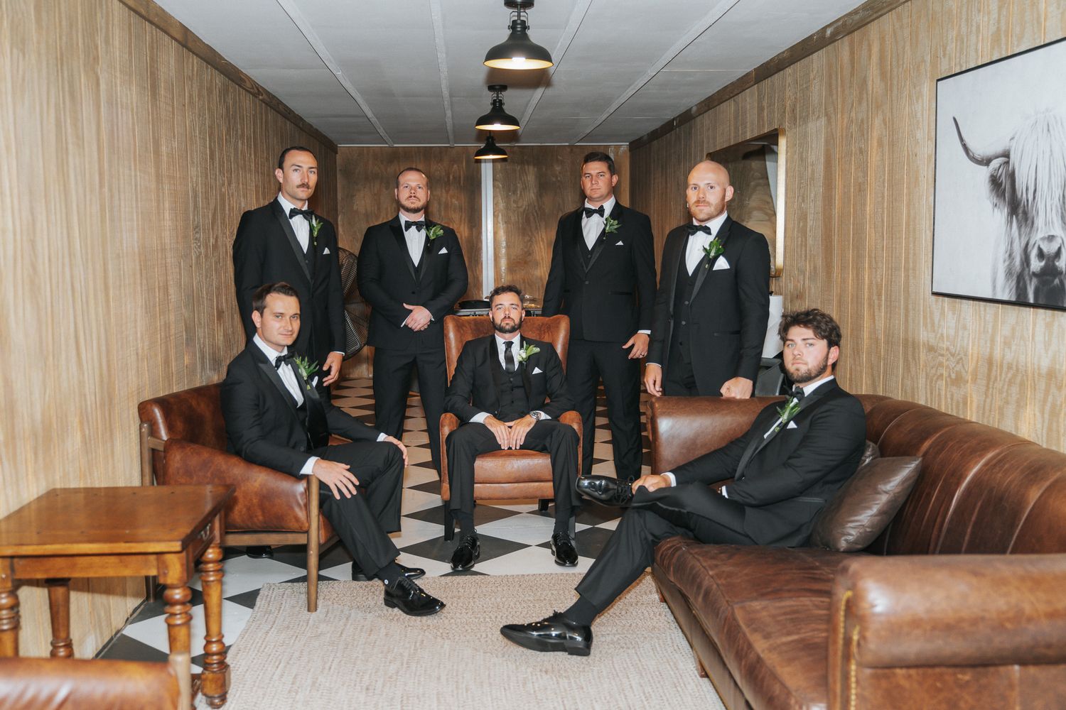 Groomsmen in black tuxedos with bow ties pose in a rustic room with leather sofas for a wedding photo.