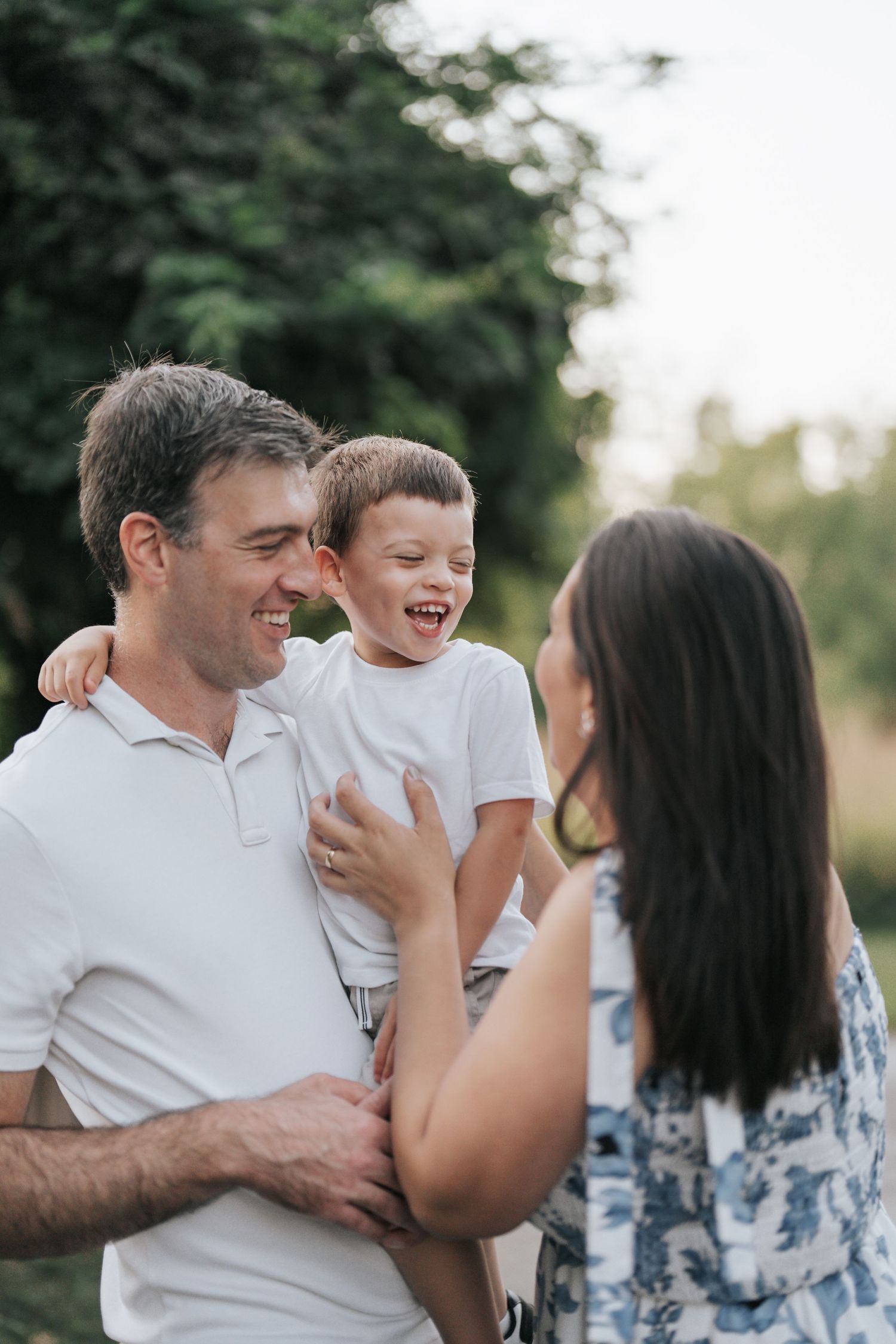 Happy family moment outdoors with father holding laughing son while mother smiles at him in a green park setting.