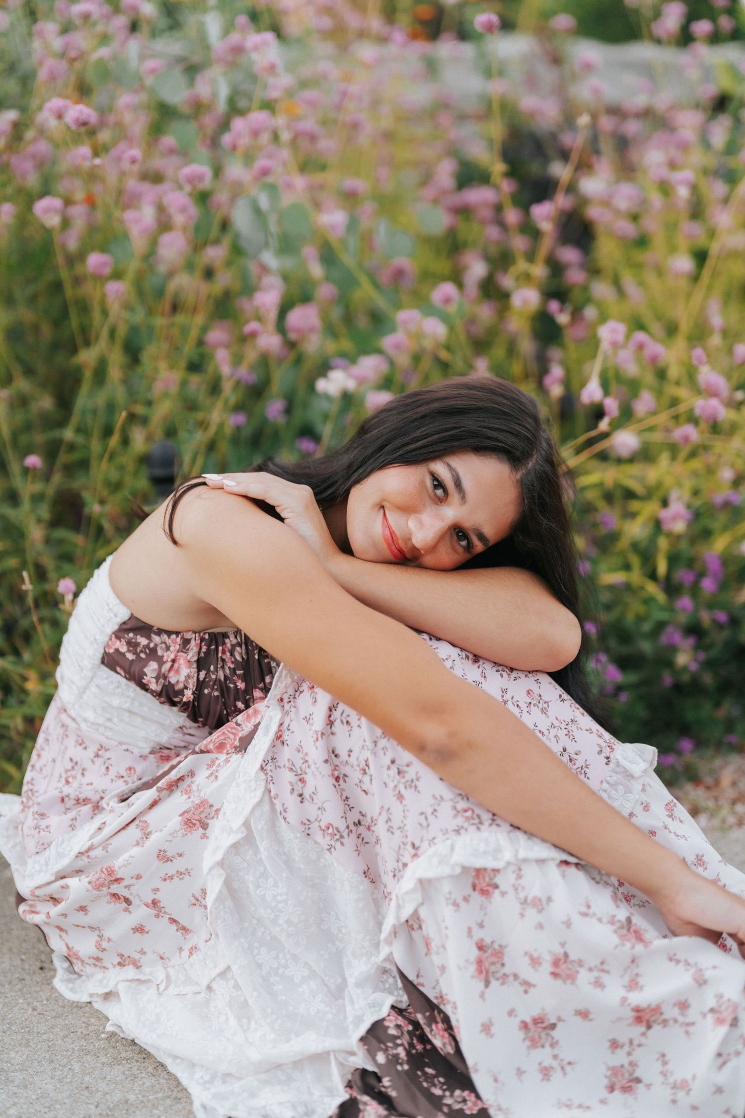 Young woman in floral dress sitting among wildflowers, smiling softly at camera.