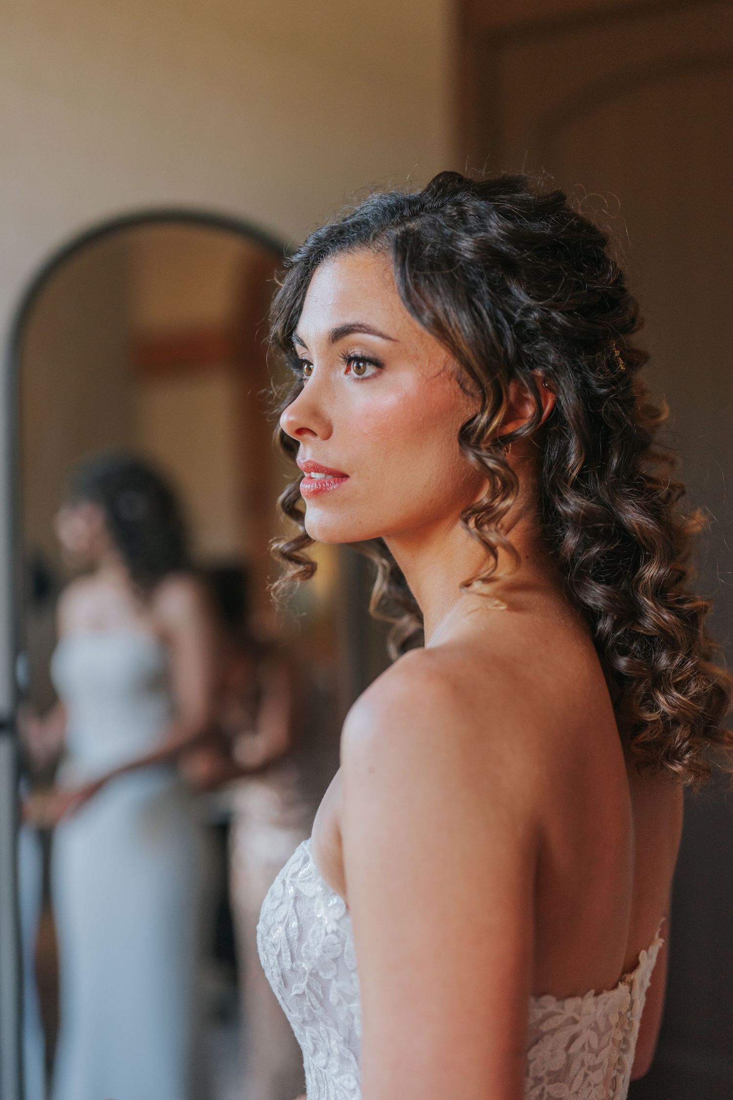 Elegant bride with curly dark hair in a lace strapless gown gazes thoughtfully in a softly lit room.