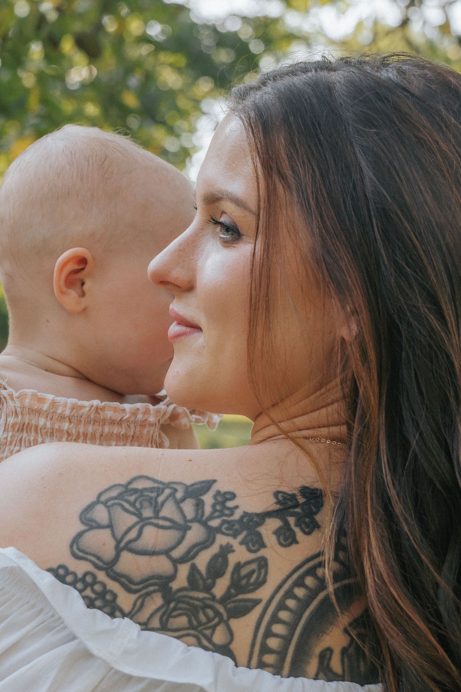A tattooed mother with long dark hair smiles while holding her baby outdoors, with a floral tattoo visible on her back.