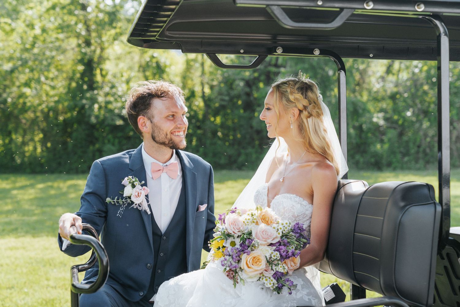 Bride and groom smile at each other while seated on a golf cart in a sunny green landscape.
