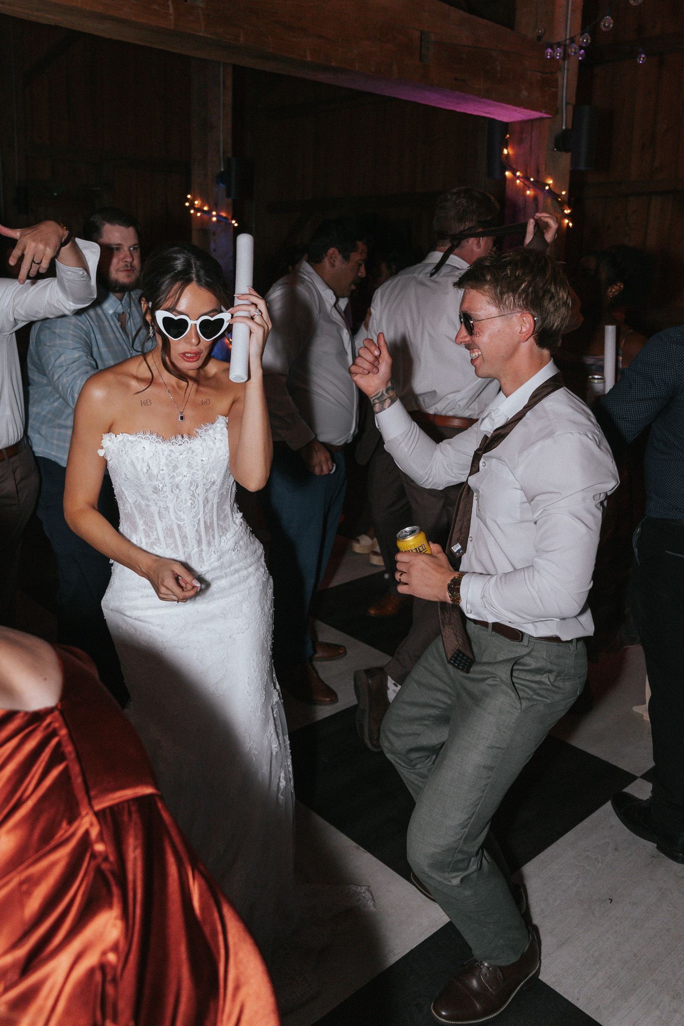 Bride in white gown and groom in suspenders dance joyfully with guests at a lively wedding reception.